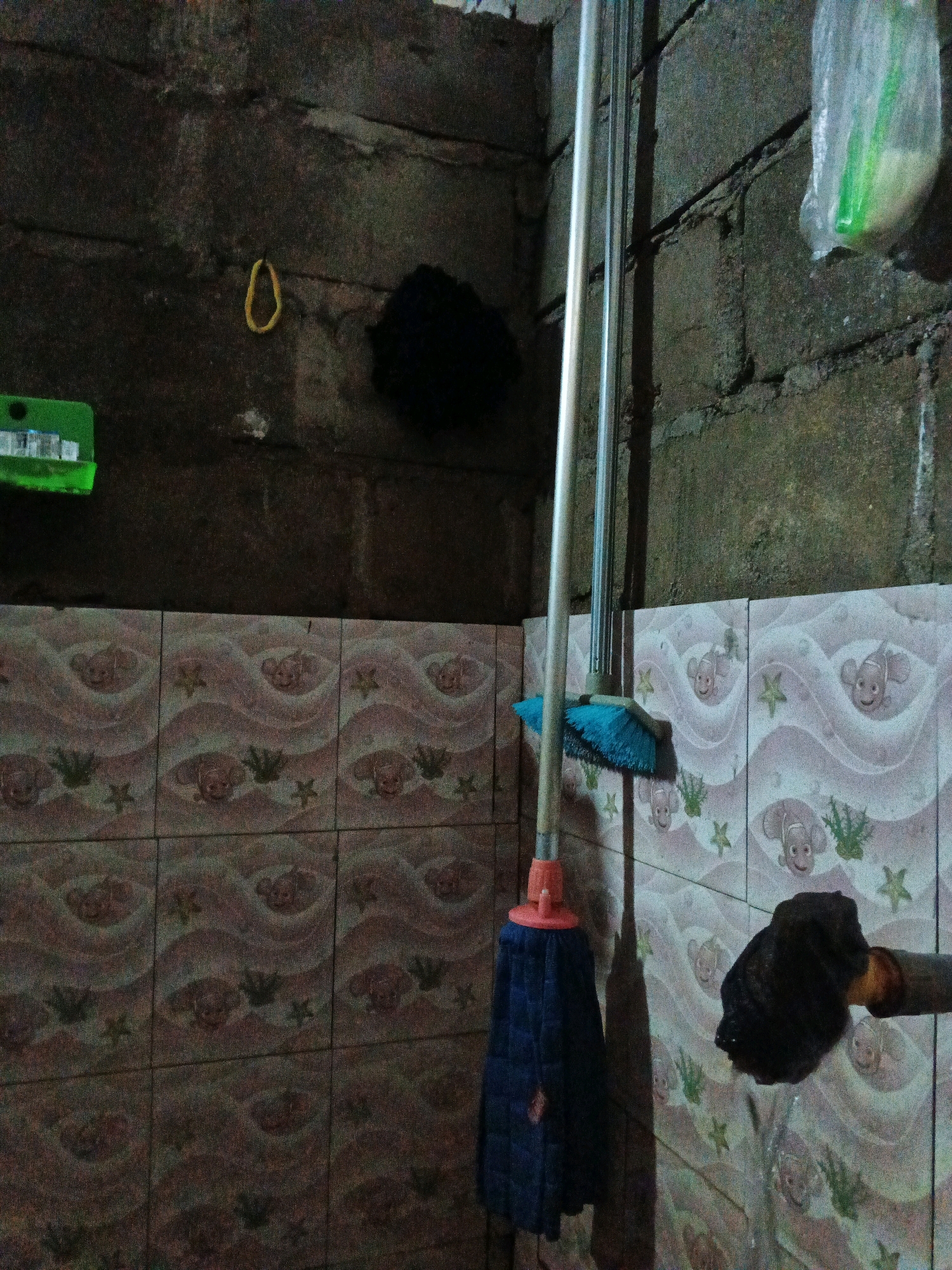 The image depicts a dimly lit, cluttered indoor space, likely a bathroom or utility area, in Brangan Mulya, Indonesia. The walls are constructed of rough concrete blocks, with some areas showing signs of moisture or moss growth. On the left, a green plastic holder with what appears to be cleaning supplies is mounted on the wall. A yellow loop, possibly a handle or hook, hangs on the wall above it. To the right of this, a dark, fuzzy object is attached to the wall.
A prominent object in the foreground is a blue mop with an orange handle, positioned vertically. Next to it, a grey metal pole, likely the handle for another cleaning tool, leans against the wall. The lower portion of the scene reveals tiled walls with a repeating pattern of light pink waves, cartoon fish, and green seaweed or plant motifs.
To the far right, a plastic bag hangs, containing a green object, possibly a sponge or soap. Below this, a dark, textured object, resembling a wet rag or sponge, is being squeezed or held, with a small amount of water appearing to drip from it. The overall impression is one of a functional, if somewhat unkempt, domestic environment.