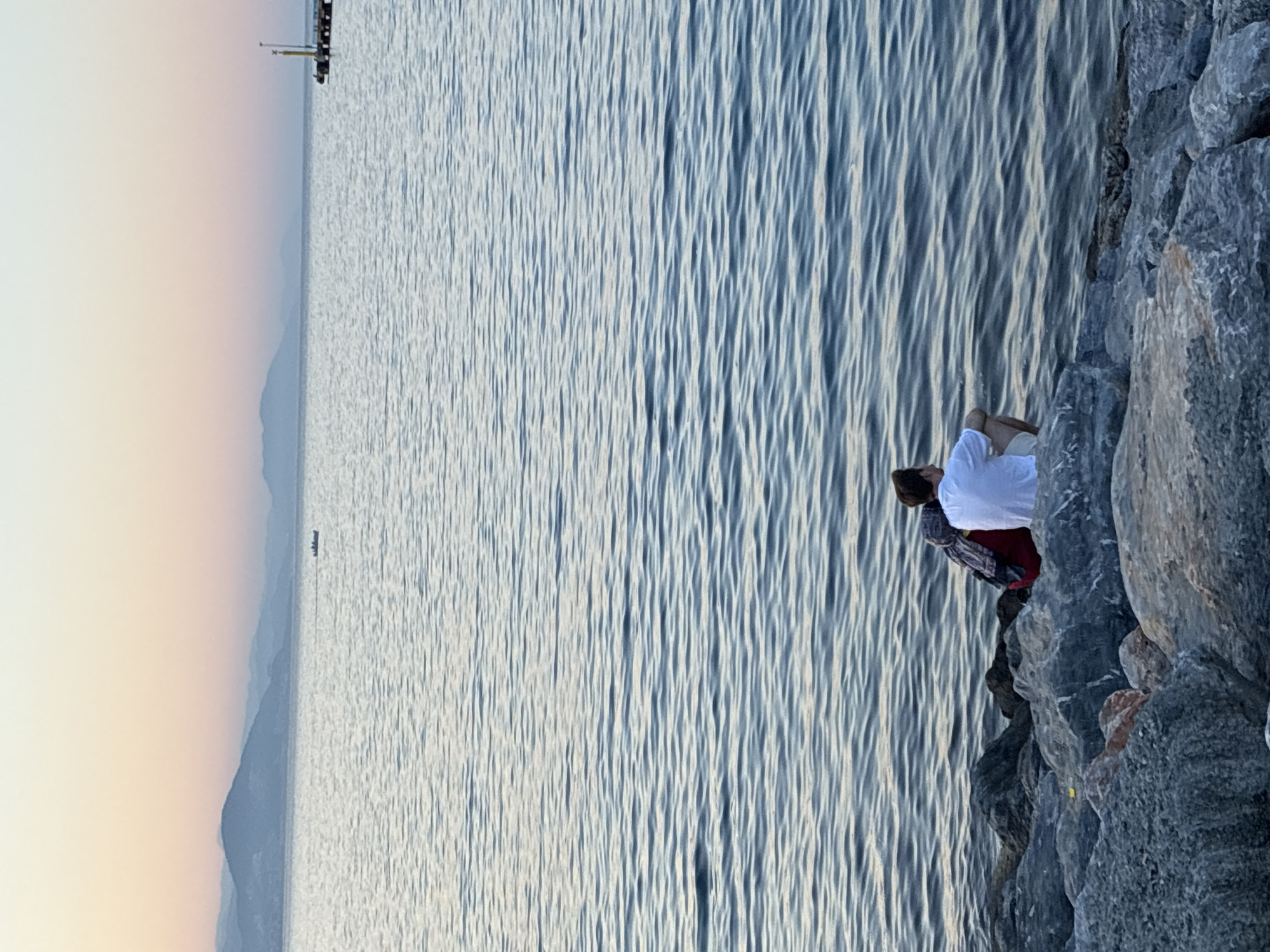 The image shows a wide shot of the sea at dusk, with mountains in the distance. In the foreground, two people are sitting on a rocky shore. One person is wearing a white t-shirt and beige shorts, facing away from the camera. The other person, to their left, is wearing a dark patterned shawl over a red garment and is also facing away from the camera. The sea is calm with small ripples. A small boat is visible in the distance, and a pier with a yellow structure is on the far right of the horizon. The sky is a gradient of pale pink and blue, indicating sunset. This scene is consistent with a location along the coast of Alanya, Turkey, likely viewed from a promenade or a rocky beach area.
