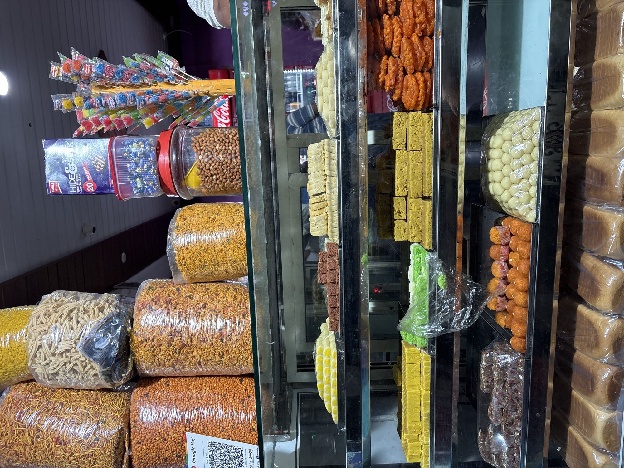 Here's a description of the image:
**Scene:** A street-side storefront in Kamalapura, India. The shop features a display of various snacks, sweets, and baked goods arranged on shelves behind a glass partition.
**Objects:**
* **Left side of display:**
* Packages of Indian "namkeen" (savory snacks), like sev, gathiya, and chivda, are stacked in plastic bags.
* Jars filled with peanuts and other small snacks
* Boxes of Parle Hide & Seek biscuits.
* Lollipops
* **Right side of display (sweets):**
* Jalebi (a sweet, fried dough).
* Burfi (Indian milk-based sweets) in multiple colors
* Laddu (ball-shaped sweets)
* Stacks of sliced bread.
**Environment:**
* The display is likely situated in a brightly lit shop, visible through the glass front.
* The storefront is likely in a busy commercial area, as evidenced by the variety of goods on offer.