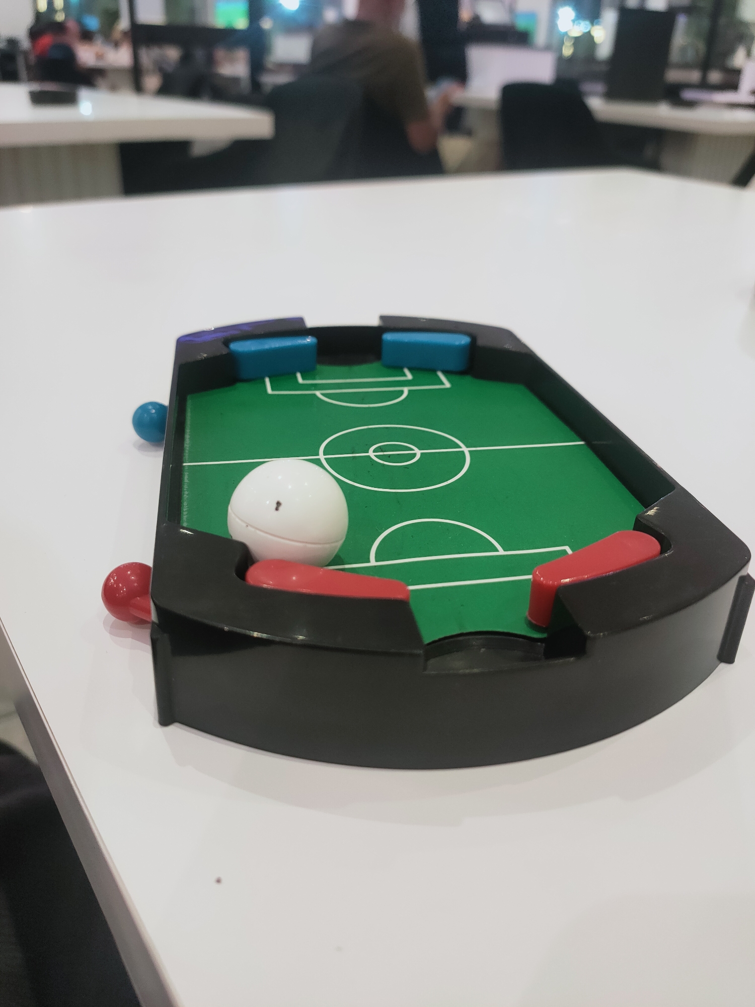 A close-up, eye-level, medium shot shows a toy foosball table with a green field and white lines, situated on a white table. The table is black with red and blue players. A white ball is in the center of the field. In the background, a blurred office environment is visible with people and computers. The location is identified as Cebalat, Tunisia.