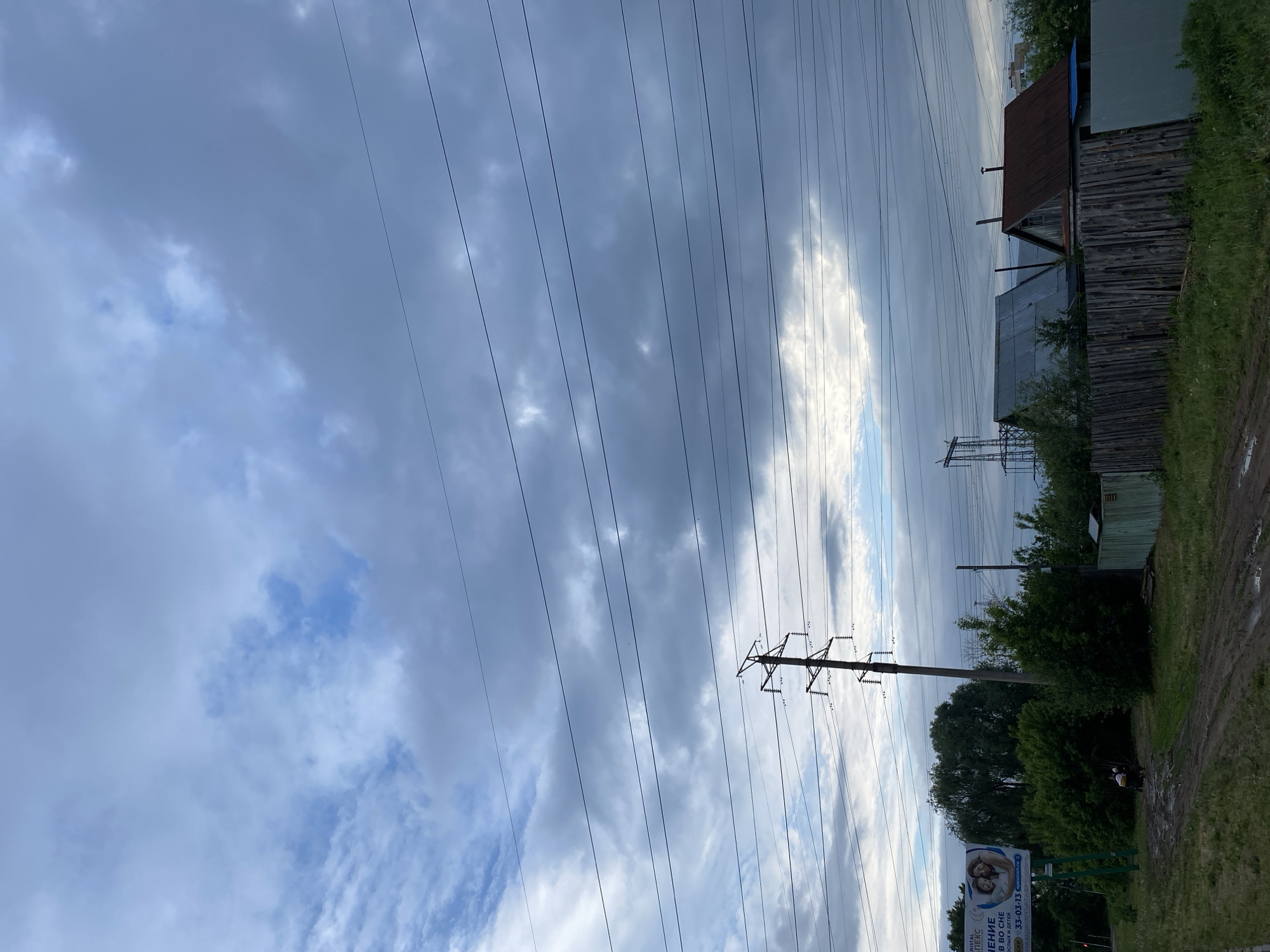 The image depicts an overcast scene in Chaikovsky, Russia, characterized by a low, heavy cloud cover. Power lines dominate the view, stretching across the sky. To the right of the frame is a wooden fence and several buildings, suggesting a residential or semi-rural area. Vegetation is present, including trees and grass, possibly along a roadside. In the lower right corner, a muddy path is visible. In the foreground there is a billboard with Russian text and a phone number, "33-03-13" partially visible. In the middle ground, a pylon supports the power lines.