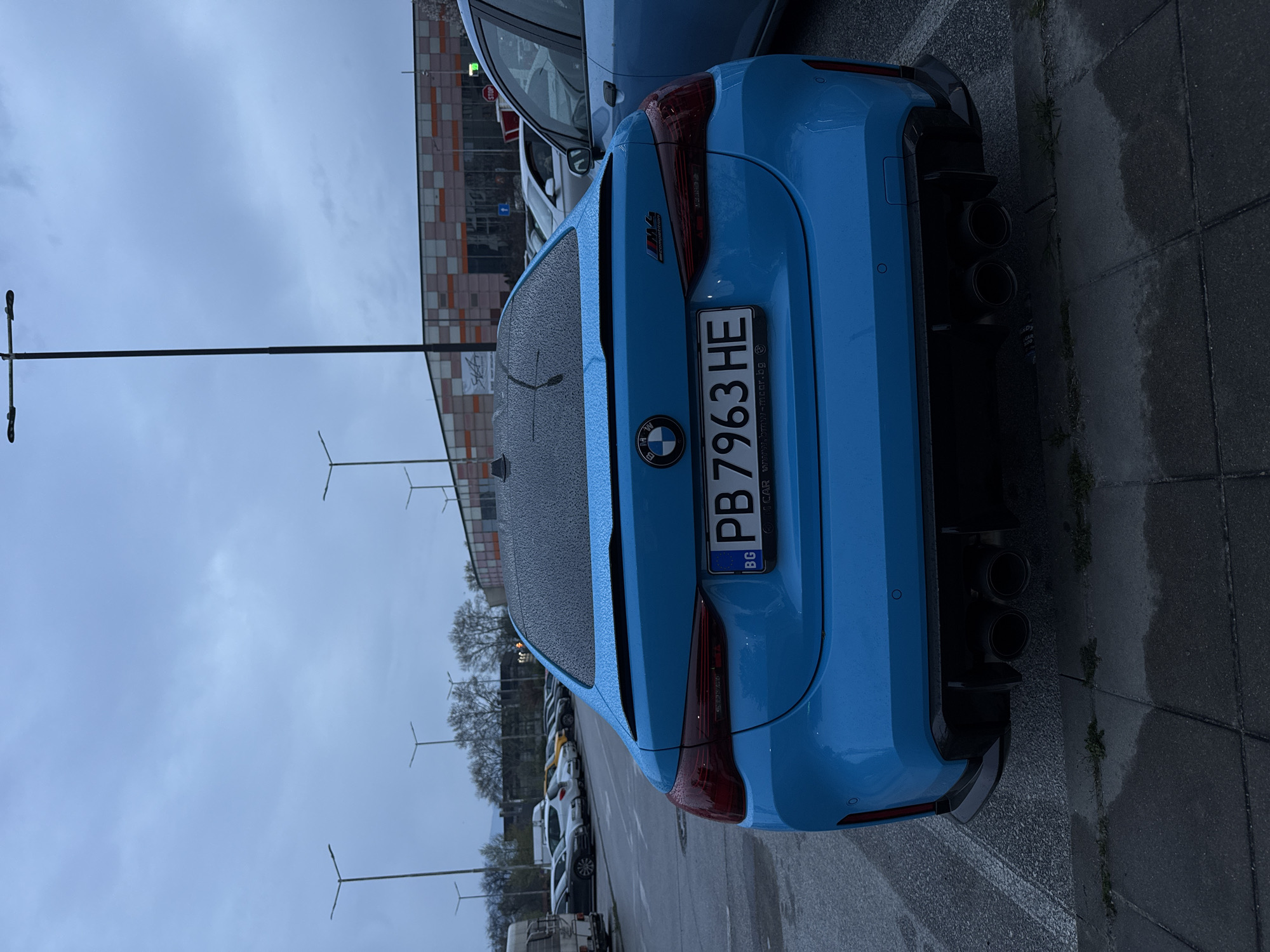 A light blue BMW M4 Competition with Bulgarian license plate PB 7963 HE is parked on the side of a street, likely near a commercial or residential area in Plovdiv, Bulgaria. The car is viewed from the rear, showing its quad exhaust system and rear lights. It's a cloudy day, and there are water droplets on the car's roof. Other parked cars can be seen in the background. Street lights are also visible in the background. The ground is wet.