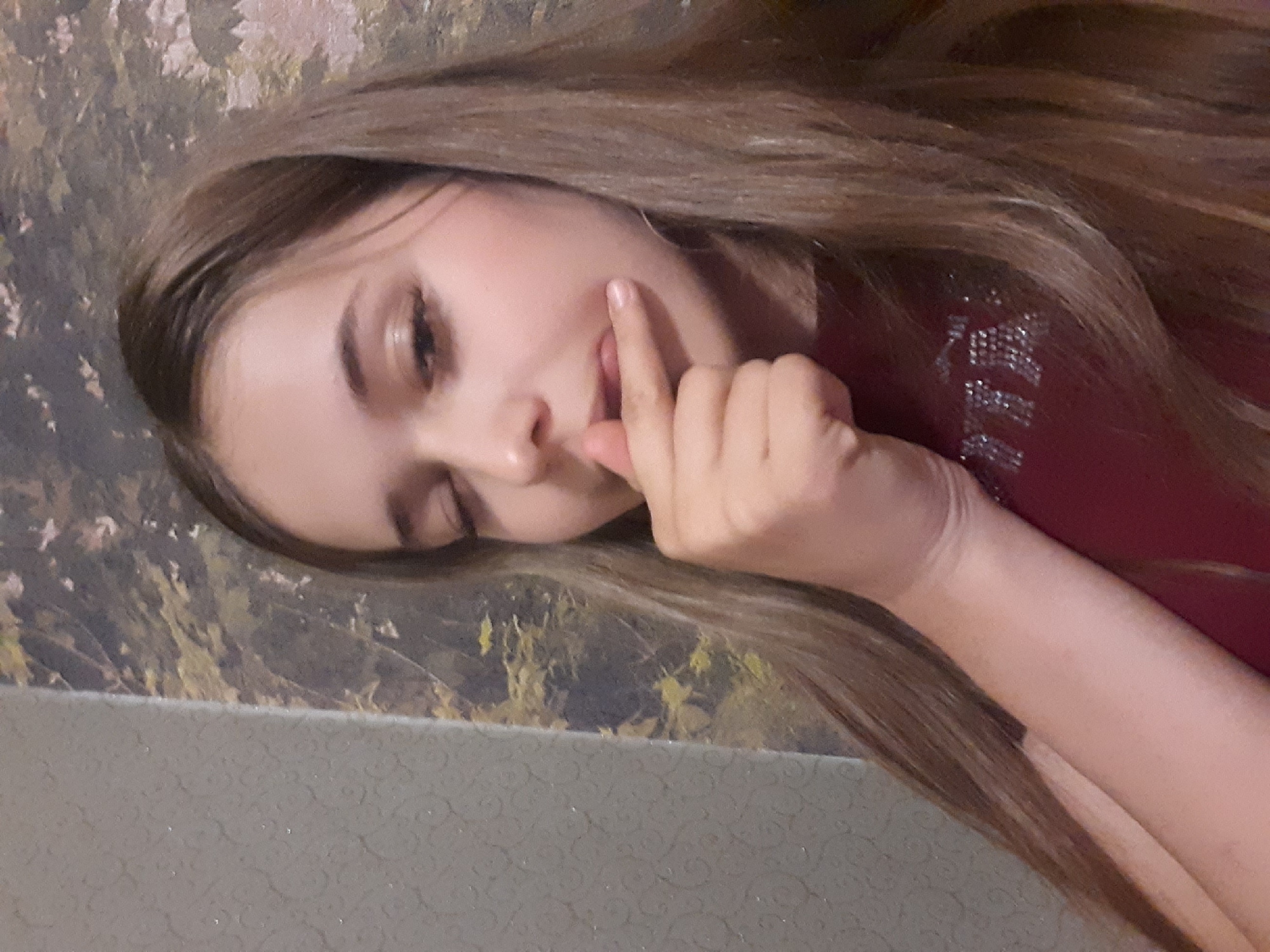 In an indoor setting with textured wallpaper in the background, a young female with long, light brown hair is pictured. She is looking down and to her right, with her right hand touching her lips. She is wearing a dark red top that has some sparkling text on it. The lighting is somewhat dim.