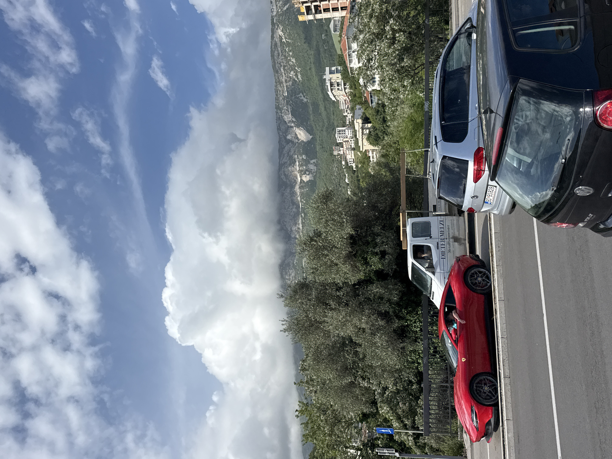 A vertically oriented photograph depicts a street scene in Boreti, Montenegro. On the right, a paved road with painted white lines is lined with parked vehicles: a black hatchback, a silver SUV, a white van, and a red sports car. The red car's driver's side window is open, revealing a person's arm. Adjacent to the road is a green area with trees and foliage. Beyond this, a hillside ascends, dotted with buildings and vegetation. To the left, a dramatic sky filled with blue sky and large white clouds dominates the scene.