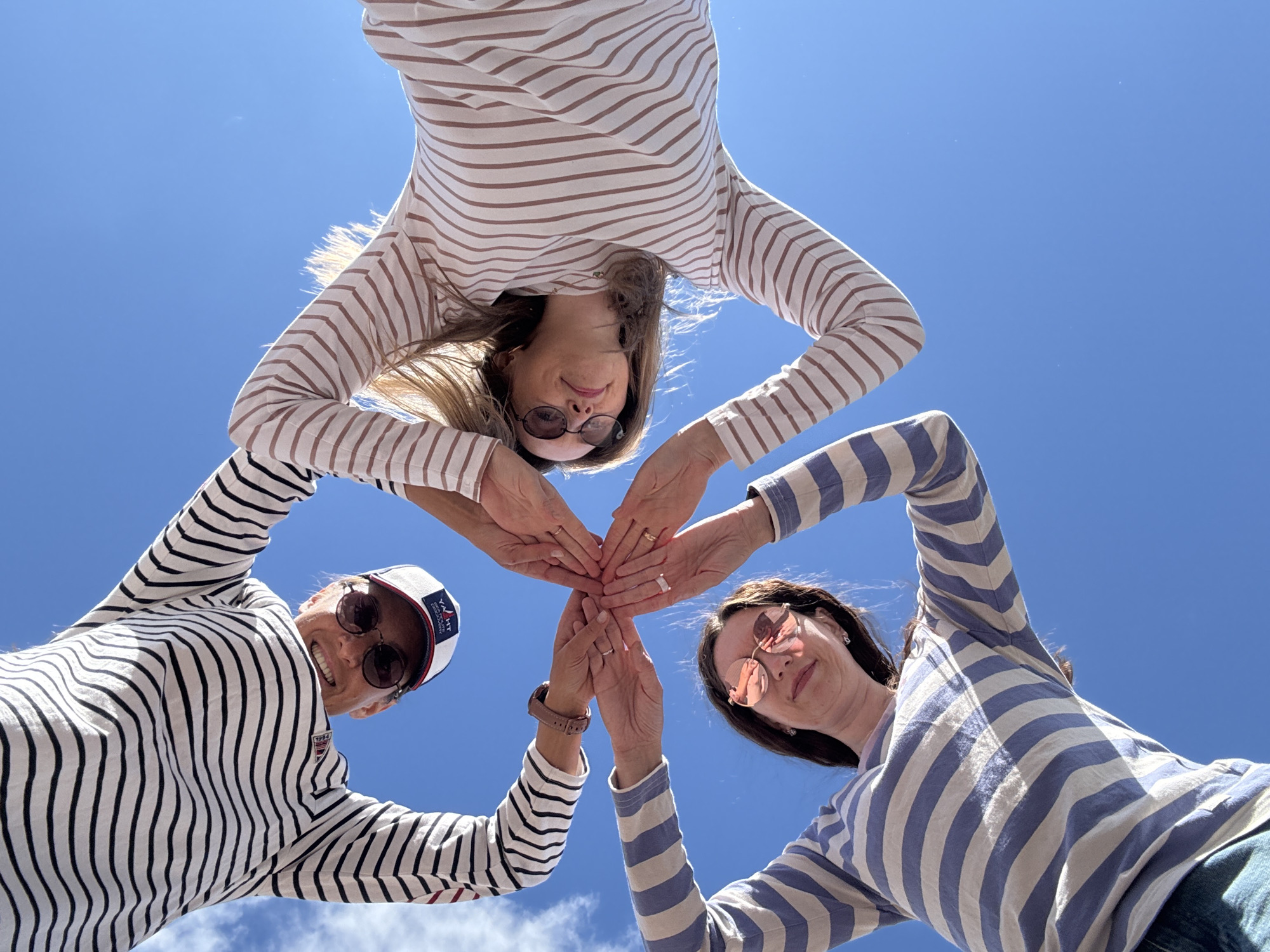 In an outdoor, low-angle shot against a bright blue sky with scattered clouds, three women wearing striped long-sleeved shirts are gathered together. They are in Chaikovsky, Russia, with their hands clasped in the center, forming a circle. The woman on the left wears a black and white striped shirt and a white baseball cap with a logo. The woman on the right has a beige and brown striped shirt and blonde hair. The woman at the bottom wears a blue and white striped shirt and round pink-tinted sunglasses. The scene captures a moment of unity and connection.