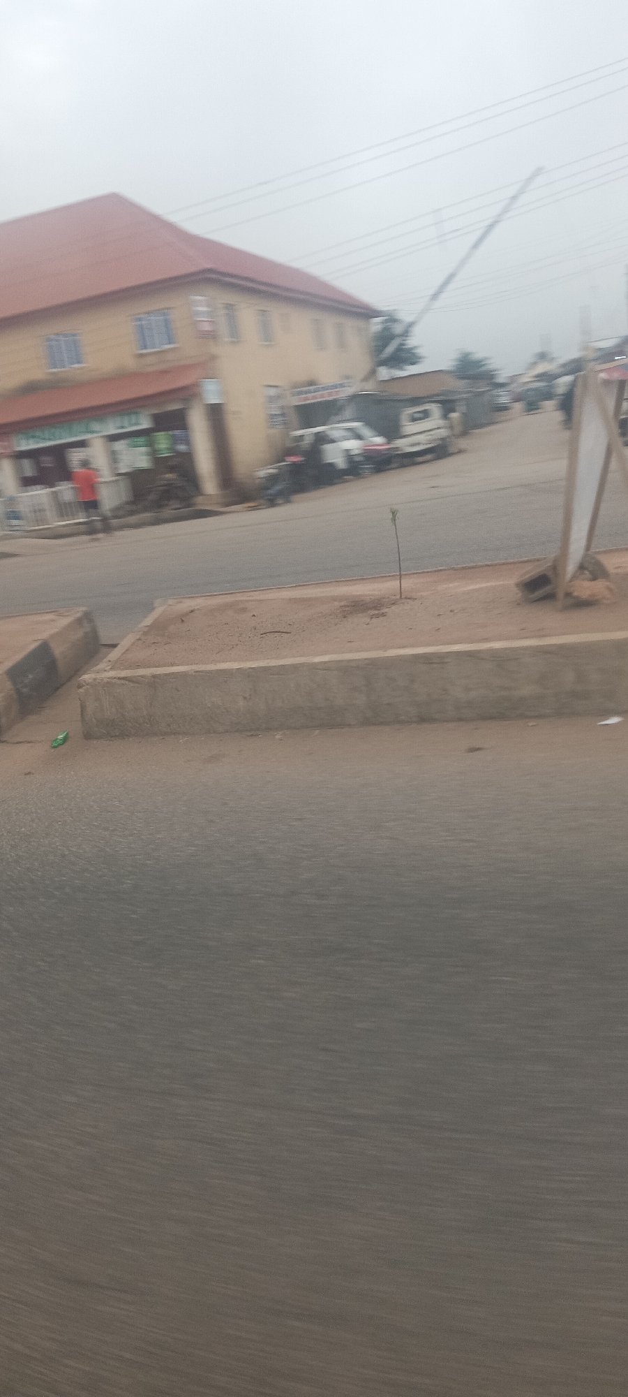 This image captures a street scene in Jos, Nigeria, under an overcast sky, suggesting diffused light. The prominent subject is a two-story cream-colored building with a reddish-brown hip roof on the left side of the frame. The ground floor of this building houses businesses, indicated by a visible sign that reads "PHARMACY LTD." Another sign on the same building appears to have "MANAGEMENT" written on it.
A person wearing a red shirt is standing near the ground-floor shops. The paved road stretches into the distance, where several vehicles, including a white van, are parked or moving. Power lines are visible against the cloudy sky above the street.
In the midground, a concrete median or barrier separates the main road from a patch of reddish-brown earth. A small, slender sapling is planted in this earth, and further to the right, a white signboard on a wooden stand is partially visible. In the immediate foreground, a dark gray paved road surface is seen, with another concrete barrier running along the bottom edge. A small piece of green litter lies near the concrete barrier on the left. The scene depicts a typical commercial or residential street, bustling with daily activity.