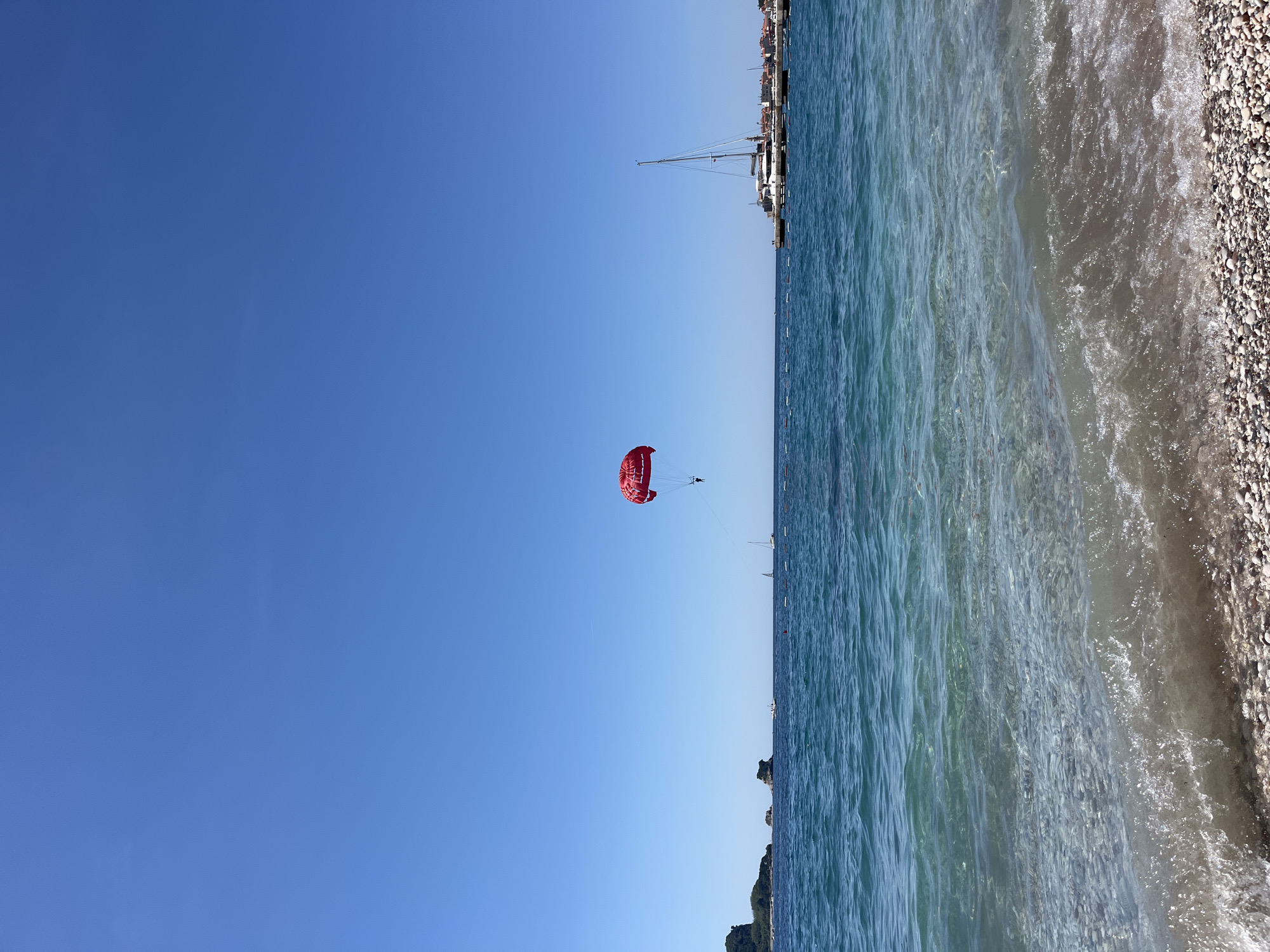 The image depicts a coastal scene in Budva, Montenegro, featuring a bright blue sky and the Adriatic Sea. A red parasail with a person suspended beneath it is visible in the center of the frame. The water transitions from deep blue to lighter shades near the shore, where a pebble beach is present. In the background, there are boats docked at a harbor or marina and the buildings can be seen along the coast. The scene suggests a clear, sunny day.