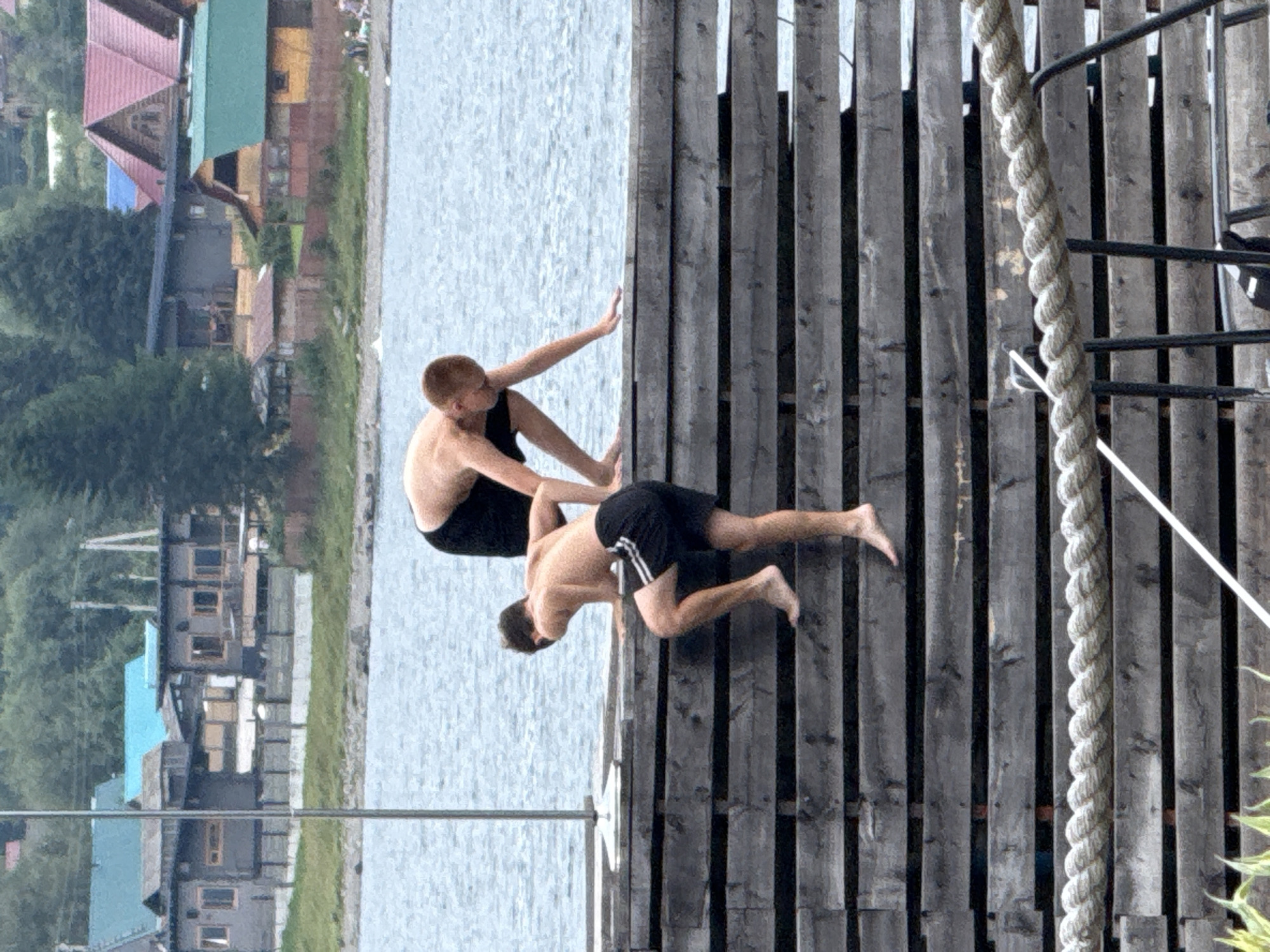 Two young men are on a wooden dock by a body of water, likely Lake Teletskoye in Artybash, Russia. The man in the foreground, wearing black swim shorts with white stripes, is climbing up the wooden slats of the dock, with his right leg extended upwards. His body is angled, with his left hand braced on the dock. The man in the background, wearing black swim shorts, is also on the dock, facing away from the camera, with his arms bent and his torso leaning forward. They are shirtless, with fair skin. The water behind them is calm and light blue. In the distance, across the water, are several buildings with various colored roofs, including blue, red, and green, nestled among green trees. The sky is overcast and the overall lighting is subdued. The wooden dock is made of wide, dark planks, arranged horizontally, with visible grain and texture. A thick, twisted rope runs horizontally across the lower portion of the image, attached to metal posts.