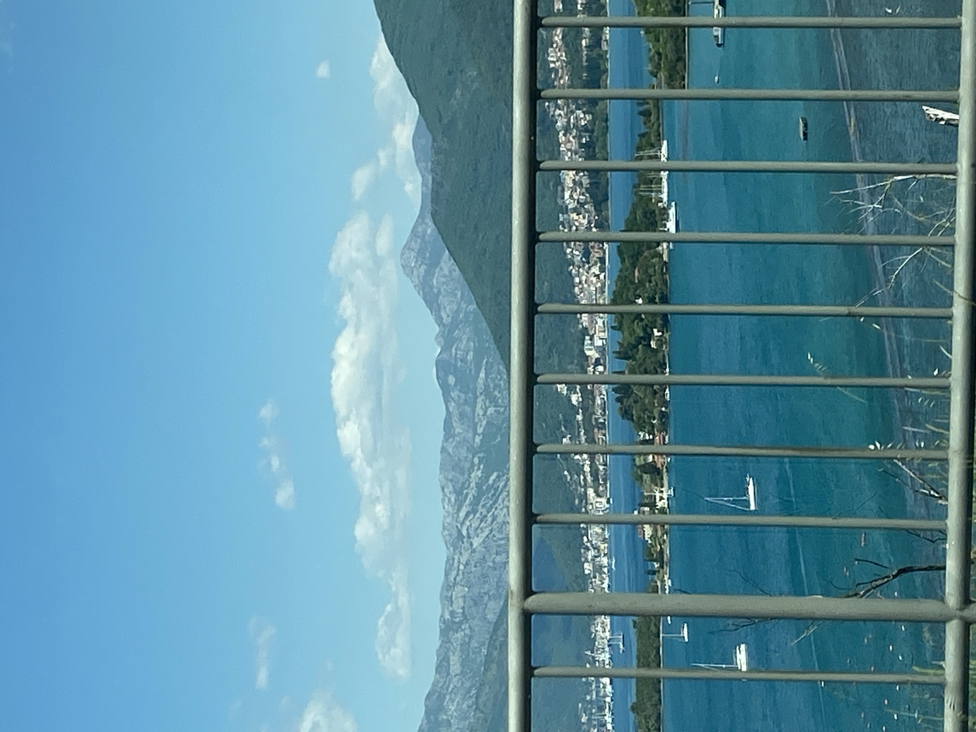 The image depicts a landscape viewed through a grey railing. In the background, there are mountains partially obscured by clouds. To the right of the mountains is a body of water with a shoreline lined with buildings and vegetation, possibly the coast of Đuraševići, Montenegro. Several sailboats are visible on the water. The sky is clear and blue with some scattered clouds.