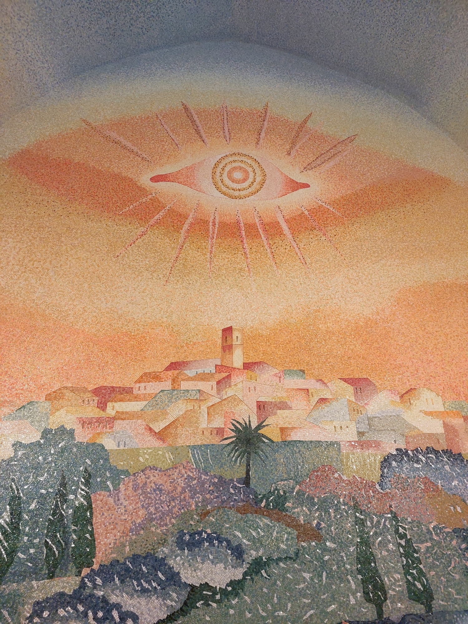 This mosaic depicts an eye-shaped sun with rays radiating outwards, set against a gradient of blue and orange sky. Below, a cluster of buildings forms a village, with a prominent bell tower. In the foreground, a landscape of trees and bushes is rendered in various shades of green, blue, and purple. This artwork is located in the Fondation Maeght in Saint-Paul-de-Vence, France.