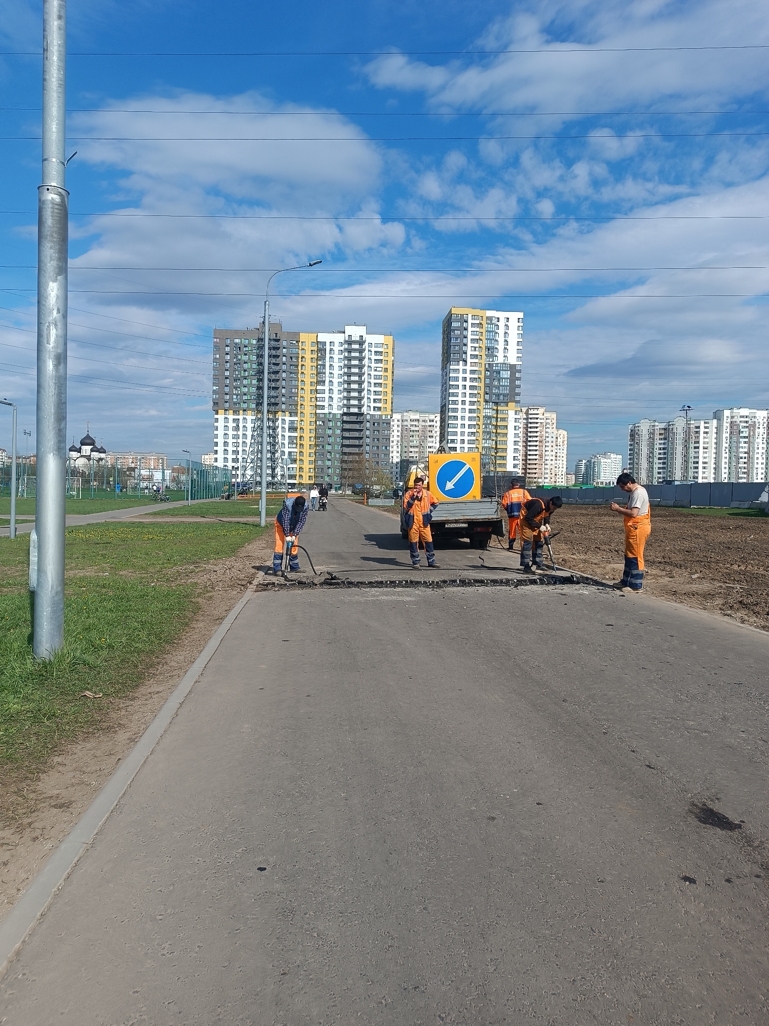 The image depicts a road repair scene in Moscow, Russia. Four workers in orange overalls are repairing the road surface. One worker is operating a jackhammer, while others are positioned around the work area. A white truck with a blue and yellow directional sign is parked nearby. The road is asphalt, and there is a grassy area on the left and bare earth on the right. In the background, there are several high-rise residential buildings and a church with a dark dome. The sky is blue with scattered clouds, and power lines stretch across the sky. A tall, metal utility pole is visible on the left side of the frame.
