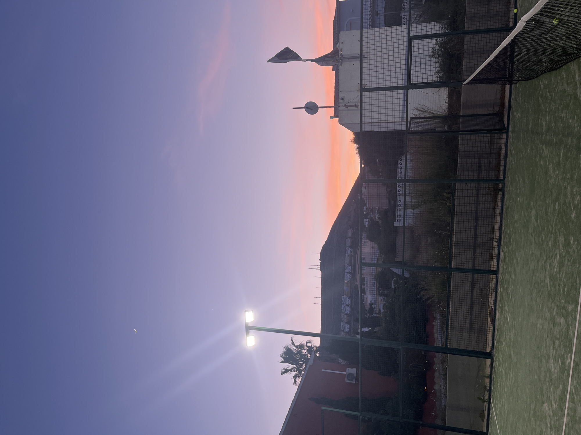 The image shows a paddle tennis court at dusk in El Poble Nou de Benitatxell / Benitachell, Spain. The sky transitions from purple to orange above a mountain with buildings. A crescent moon is visible in the dark blue sky. A court light illuminates the bottom left. The court's fence is metal mesh. A tennis ball is stuck in the net.