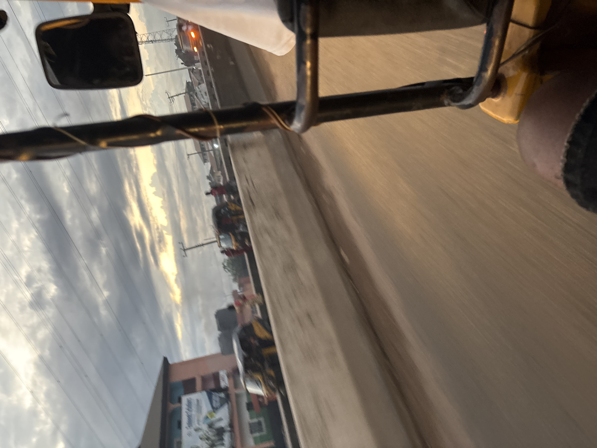 An eye-level, outward-facing shot from a moving vehicle, possibly a tuk-tuk, captures a street scene in Onitsha, Nigeria. The sky is overcast with dramatic clouds, partially illuminated by the setting sun. Power lines crisscross the sky above a low-rise building on the left, which has a billboard advertisement. In the middle ground, several vehicles, including at least two tuk-tuks and a car with its brake lights illuminated, are on a road separated by a concrete barrier. The foreground shows the leg of a person and part of the interior of the vehicle, suggesting the photographer is a passenger. The overall impression is one of a busy urban environment during twilight.