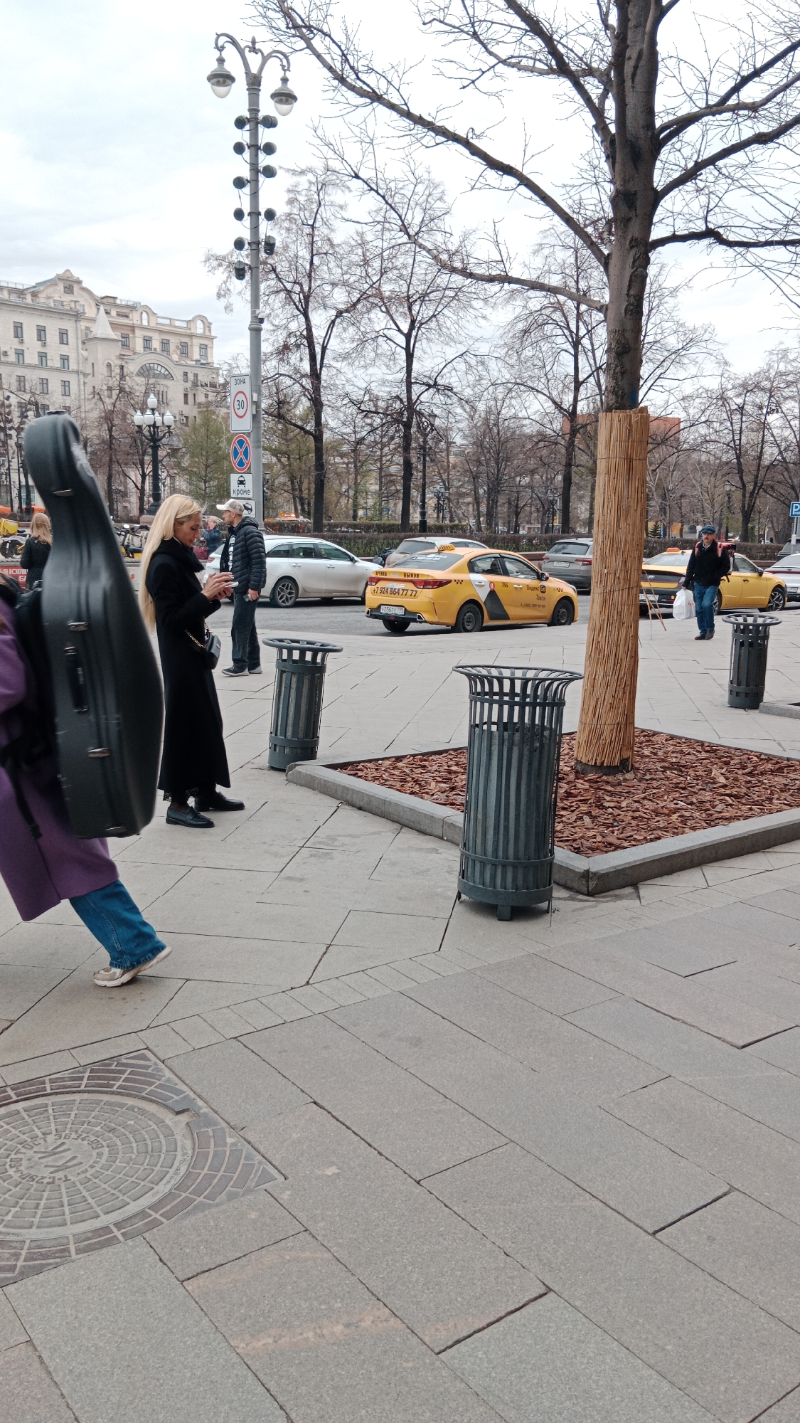 The scene is an urban environment in Urshel'skii, Russia. Several people are present: one carrying a cello case, a blonde woman in a black coat looking at a phone, and others further in the background. There are two distinctive yellow taxi cars parked on the street, with Cyrillic characters on their sides and phone numbers. Other cars are visible further down the street. On the paved walkway, there are three metal trash cans positioned around a tree that's surrounded by a square of bark mulch bordered by stone. A lamppost with several lights is visible in the background along with buildings and bare trees, suggesting it might be autumn or winter.