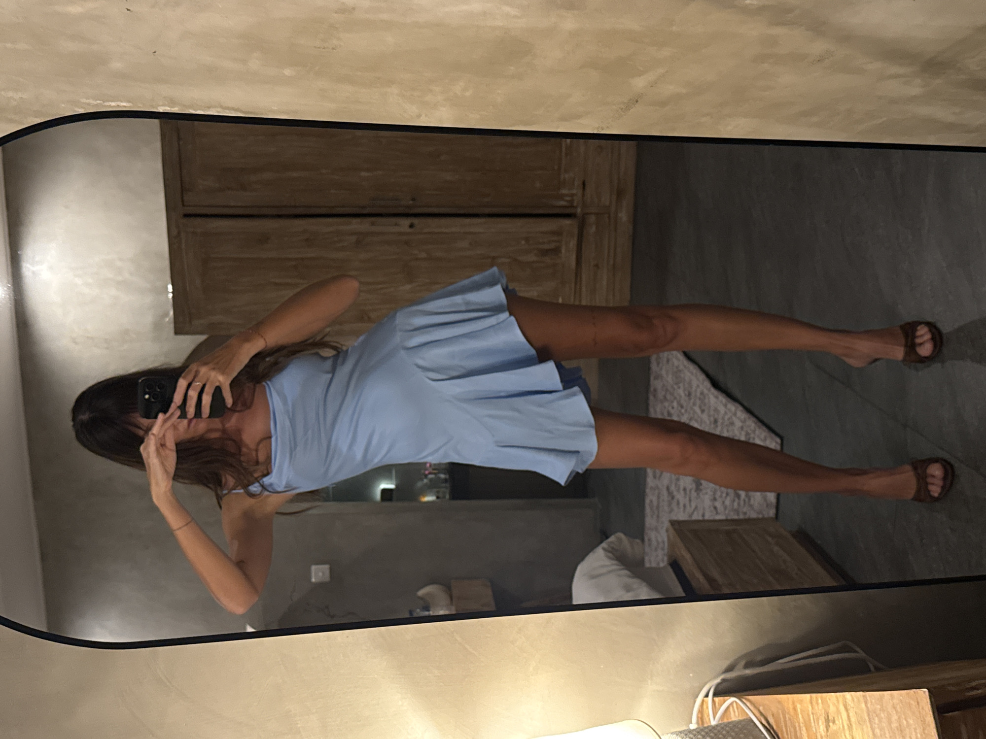A woman is standing in a room in North Kuta, Indonesia, taking a picture of herself in a mirror. The room has a minimalist design with muted tones and natural materials. The woman is wearing a light blue dress and brown sandals. The mirror is rectangular with a black frame, and it reflects a wooden cabinet and a dark gray floor. A rug is visible in the reflection. A lamp sits on a wooden desk in the foreground.