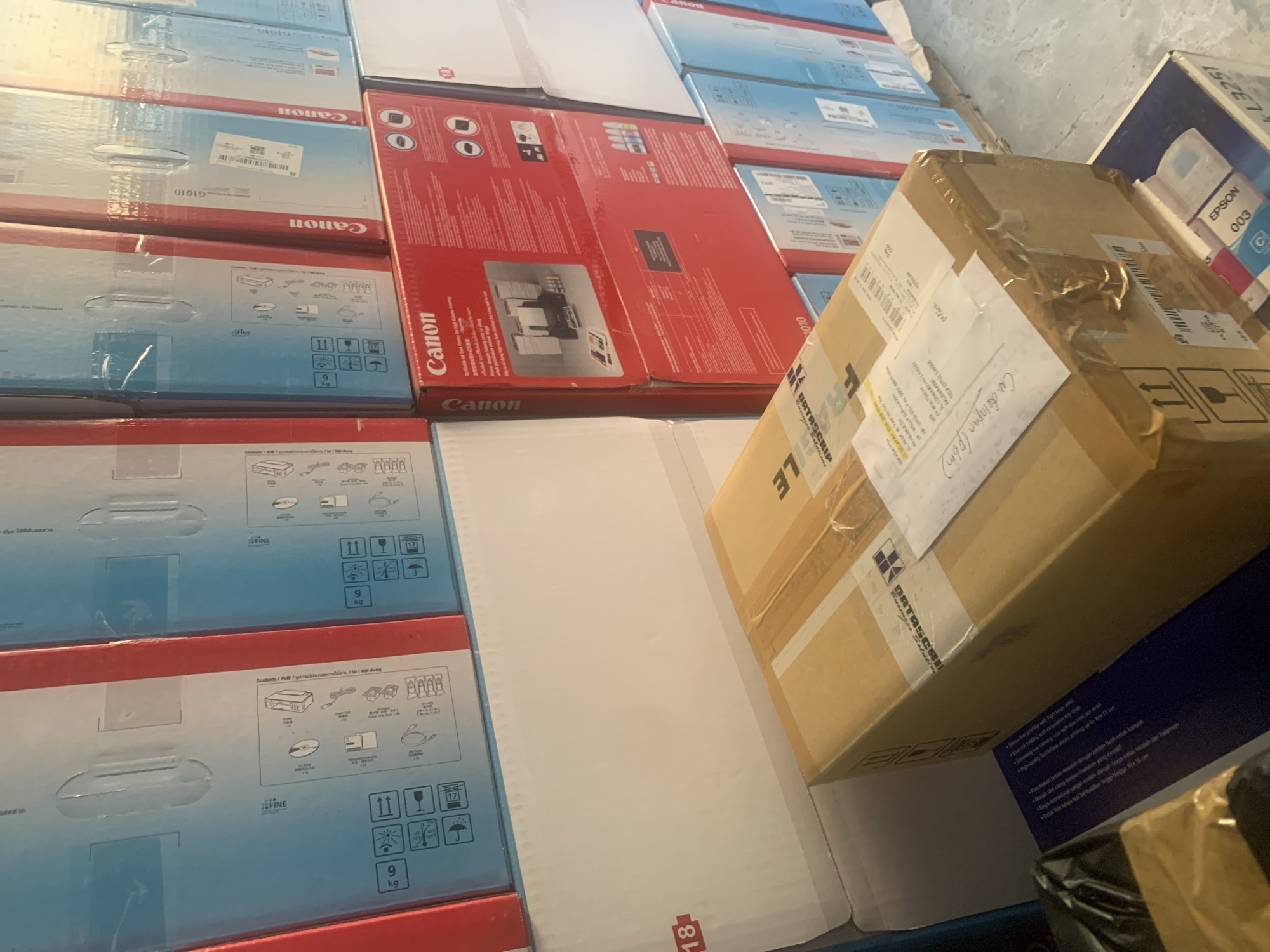 In Palembang, Indonesia, a stack of various printer boxes and a shipping package are visible. The printer boxes, primarily Canon brand, are arranged in columns and rows, with some featuring red and blue designs and Chinese characters. One prominent Canon box is bright red, displaying an image of a printer and ink cartridges. A large, white shipping box is positioned in the foreground, topped with a brown cardboard package secured with tape. This package has shipping labels and handwritten notes. Below the brown package, the edge of an Epson printer box with the model number "003" is visible. The overall scene suggests a storage or retail environment, possibly a warehouse or stockroom.