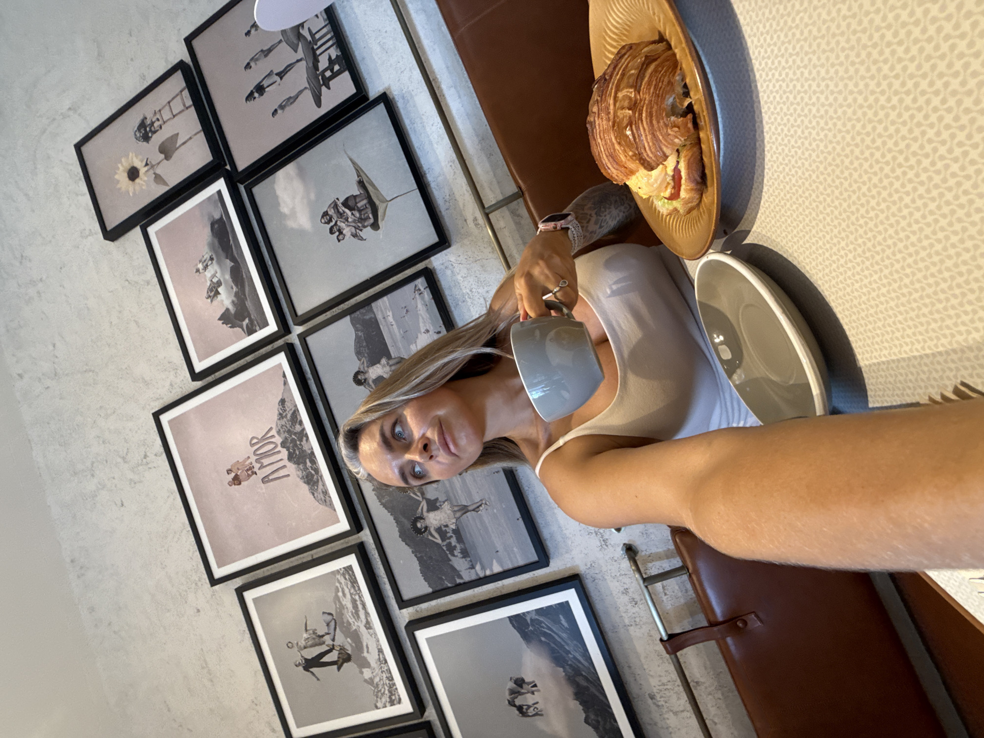 In Shanghai, China, a woman with blonde hair and blue eyes, wearing a white tank top, is seated at a table. She is holding a grey mug in her right hand and looking upwards and to her right. In front of her is a brown plate with a croissant sandwich and an empty grey bowl. Behind her, a brown leather bench is visible against a wall adorned with several framed artworks. The artworks appear to be collages or vintage-style prints depicting people in surreal or adventurous settings, some set against mountainous backdrops, and one featuring a sunflower. The overall scene suggests a cafe or restaurant setting.