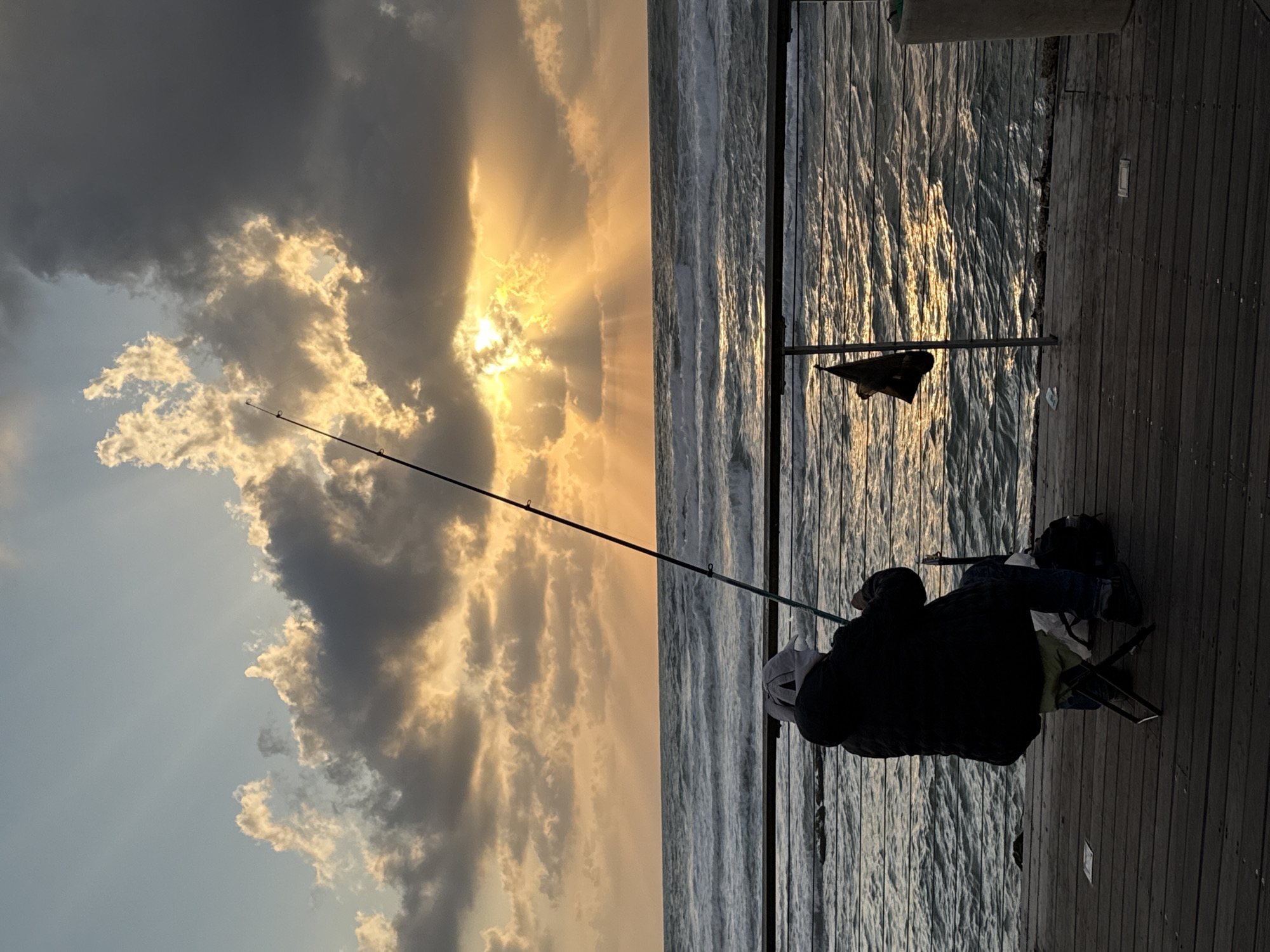 A photograph shows a person fishing on a pier or dock, likely at sunset. The person is sitting in a chair, facing the water, with a fishing rod extending out over the water. The sea is choppy with the reflection of the sun. To the left is a dramatic sky with the sun breaking through the clouds. The wooden boards of the pier run horizontally across the lower portion of the image. The picture may have been taken in the city of Tel Aviv, Israel.