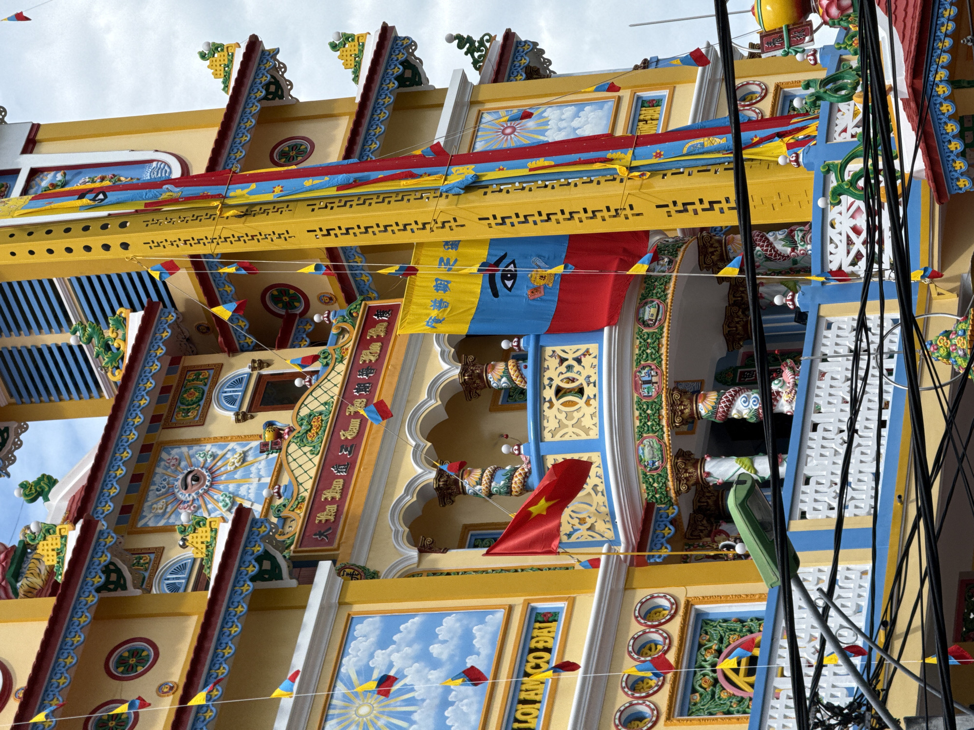 The image captures the vibrant exterior of the Long Son Pagoda in Nha Trang, Vietnam. The pagoda's facade is predominantly yellow, adorned with intricate blue and red decorative trim, small colorful flags, and large murals depicting religious scenes, including an eye surrounded by sun rays. A tall, yellow, perforated structure stands prominently on the right side of the frame, draped with colorful banners. Below the pagoda's balconies, a white railing with blue accents is visible, and numerous black electrical wires crisscross the lower part of the image, obscuring some of the lower architectural details. The sky is overcast, suggesting a cloudy day. No people are visible in the frame.