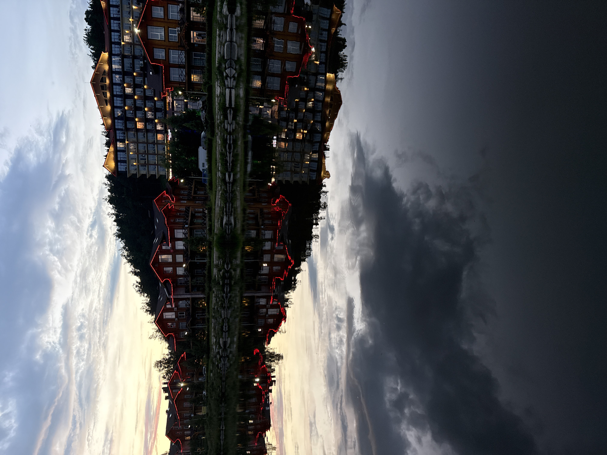 The image depicts a twilight scene at a resort in Syrostan, Russia, with buildings reflected in a body of water. The buildings, primarily wooden lodges and a larger hotel-like structure, are adorned with red lights along their roofs and edges. The sky above is a gradient of dusk colors, with clouds visible. The water in the foreground reflects the illuminated buildings and the sky, creating a symmetrical composition. The overall atmosphere is serene and suggests a peaceful evening at a vacation destination.