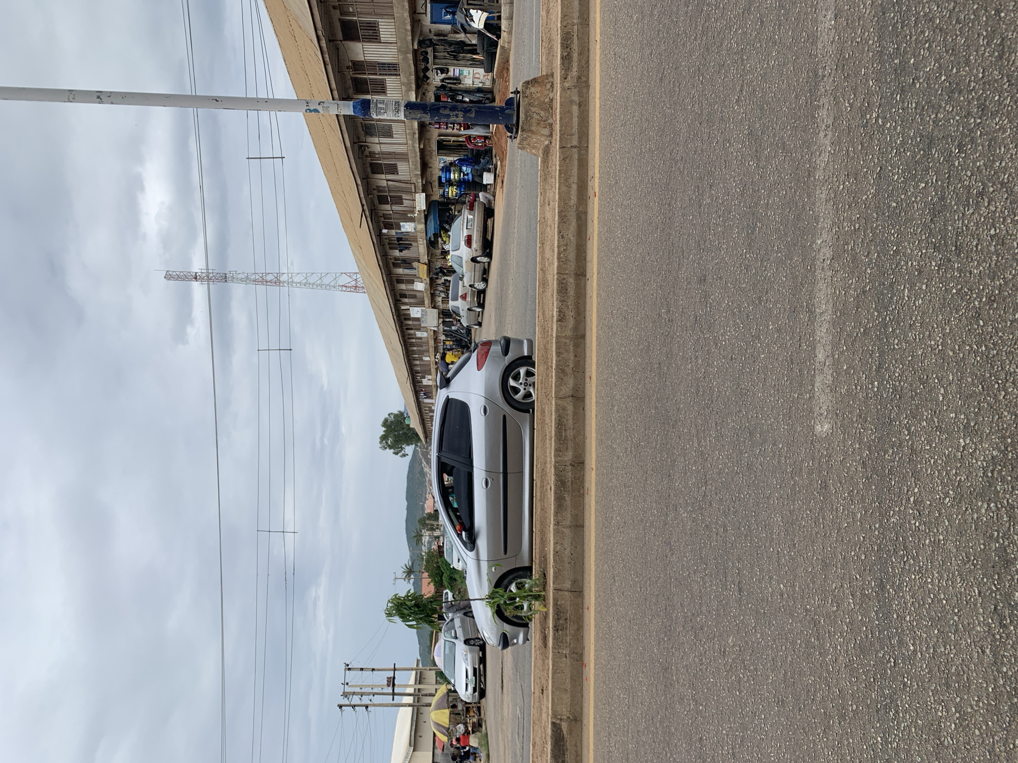 This image depicts a street scene in New Stadium, Nigeria, under an overcast sky. The foreground features an asphalt road surface with a faint white line. A concrete median separates the foreground road from a parallel parking or service lane.
Parked on the far side of the median, a silver Peugeot 206 hatchback with tinted windows and custom wheels faces left. A small, leafy plant grows directly in front of the car. Further to the left, beyond the median, a silver Toyota Corolla sedan is parked near a yellow and red striped market umbrella. Behind the umbrella, several individuals are discernible, one of whom appears to be seated under the stall.
Dominating the right side of the background is a long, two-story building with a brown corrugated metal roof. The ground floor consists of numerous open-front shops displaying merchandise such as tires and auto parts. Several individuals are present near these shops, consistent with commercial activity. The second story features balconies with metal railings. A gold/beige Honda Accord sedan is parked in front of this row of shops. A tall white utility pole with a blue base and faded posters is prominently located on the right side of the median, near the building.
In the mid-distance on the left, a cluster of wooden utility poles with multiple overhead power lines is visible. Against the cloudy sky, a tall, red and white lattice-structure communication tower stands in the upper background. Distant hills or mountains are faintly visible on the horizon towards the left. Overhead power lines also traverse the upper portion of the frame.