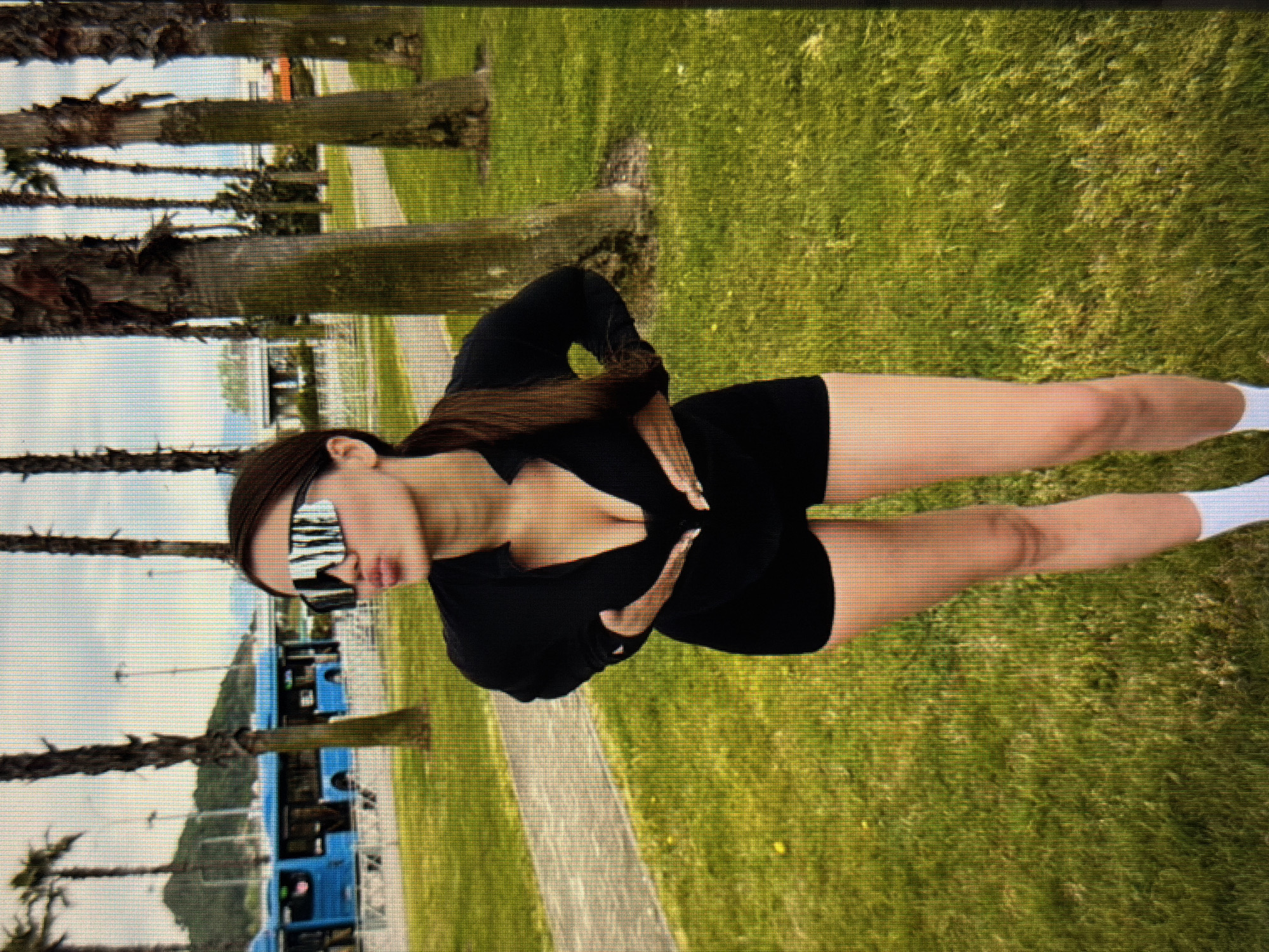 A full-body, slightly low-angle shot features a young woman with dark hair pulled back into a ponytail. She is wearing oversized, reflective sunglasses and a black, low-cut top and black shorts. Her hands are placed on her chest, and she is puckering her lips as if to kiss. She stands in a grassy area with palm trees in the background. A blue bus is visible in the distance, along with a hill and some buildings. The environment appears to be an outdoor public space.