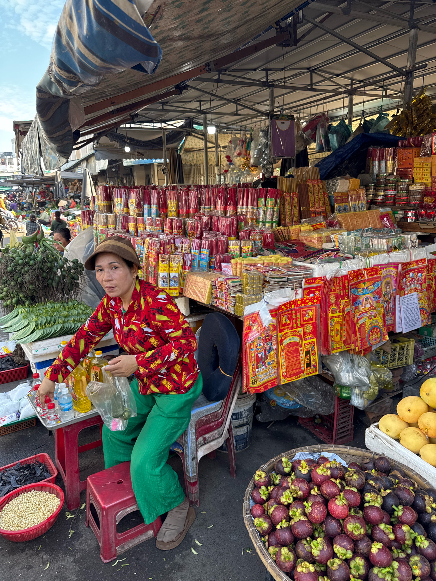 A woman wearing a brown hat, red patterned shirt, and green pants sits on a red stool in a market. She holds a clear plastic bag in her hands. In front of her is a red table with bottles of liquid and a tray with small red-capped bottles and a clear bottle. To her left, a basket of light-colored beans sits next to a red basket of dark items. In the background, bundles of green leaves and other produce are displayed. Further back, stalls are filled with various goods, including what appear to be incense sticks and religious offerings. A basket of mangosteens and some yellow fruits are visible in the foreground to the right. The scene is likely a street market in Nha Trang, Vietnam, given the presence of Vietnamese text on some of the items and the overall market atmosphere.