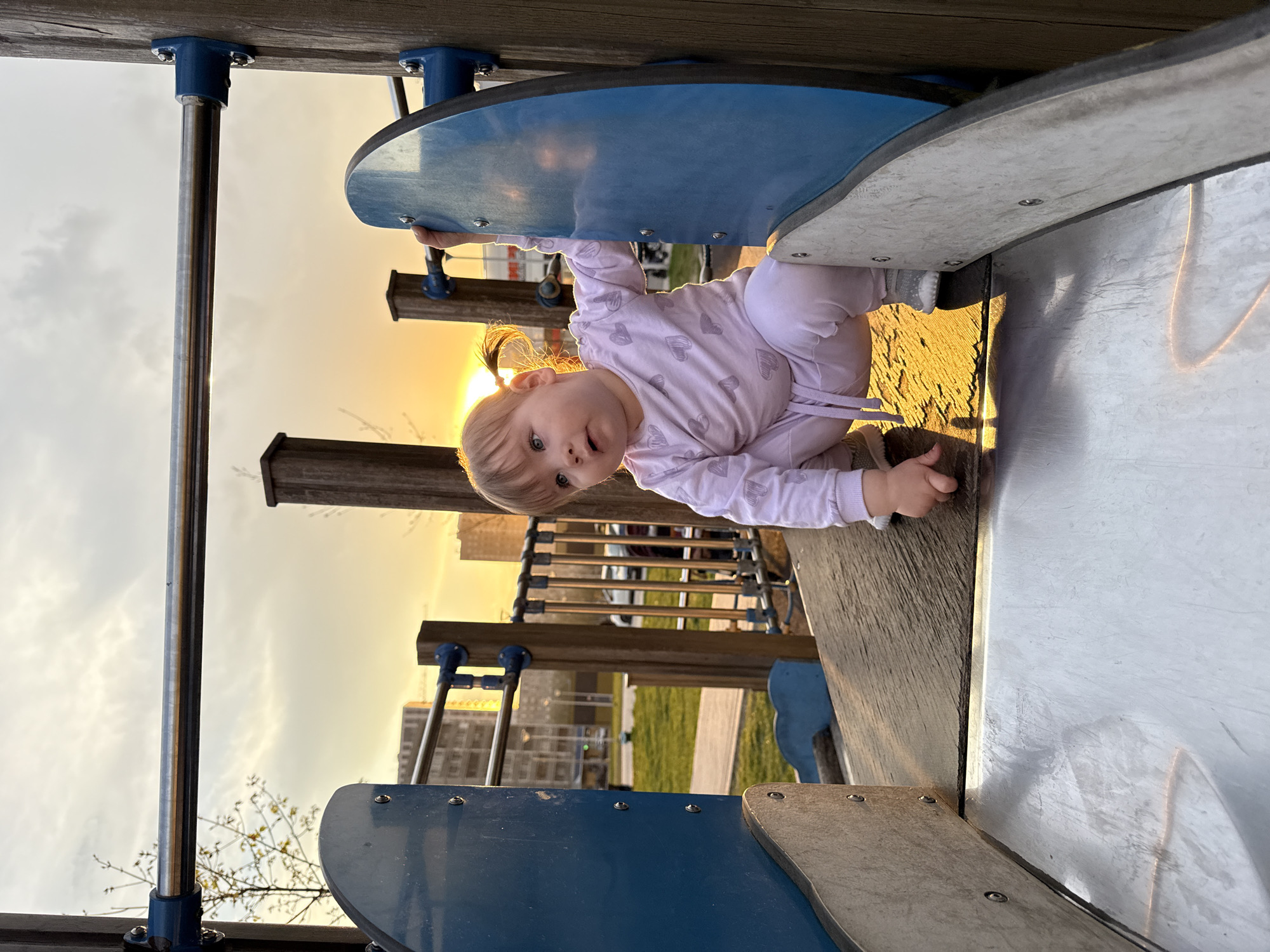 A baby is on a playground in Moscow, Russia. The baby is wearing a pink outfit with a gray heart pattern. The playground features a slide with blue and gray elements and a metal support post. The background shows a park with green grass, buildings, and a partly cloudy sky at sunset.