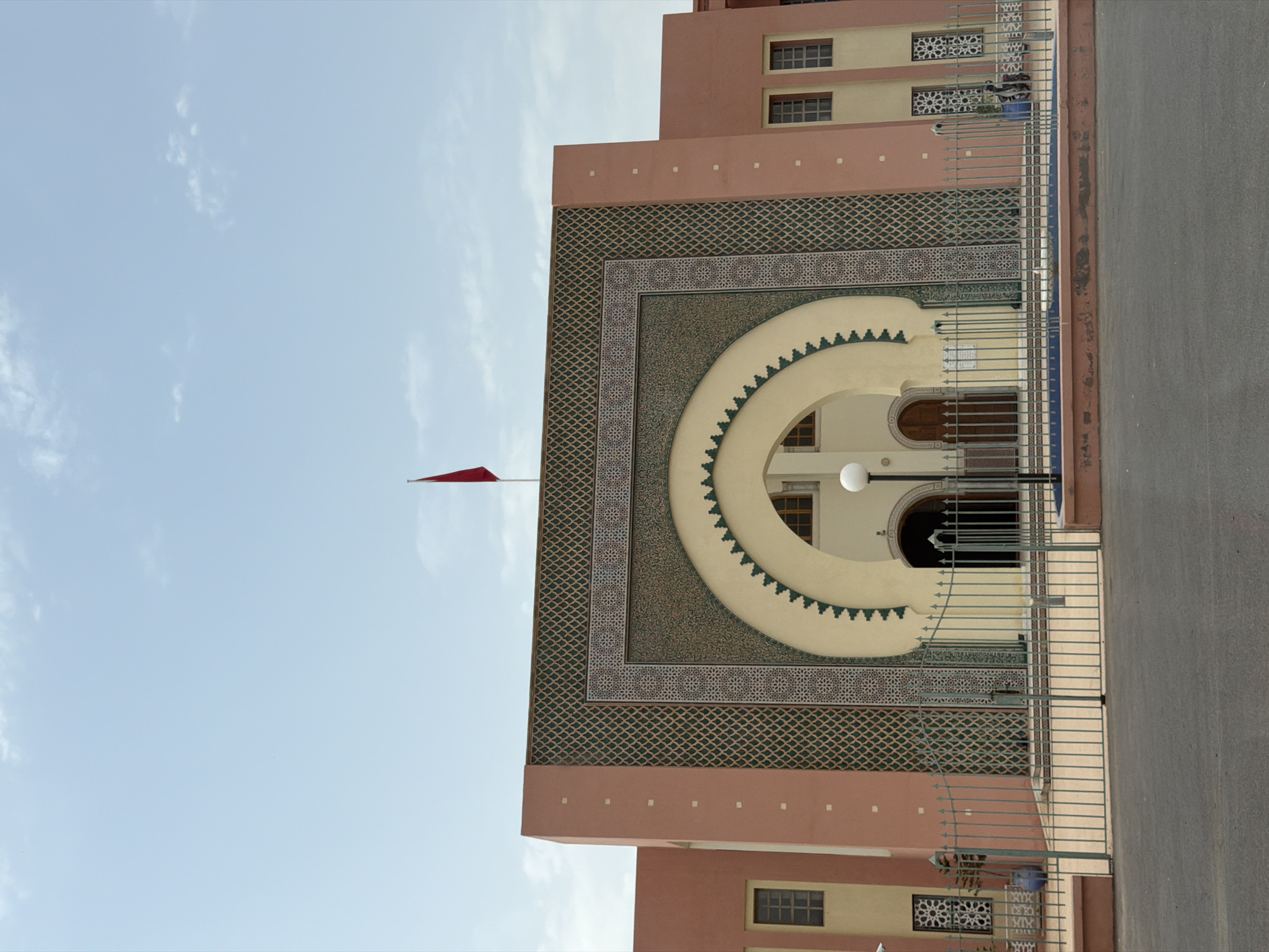 The image captures the exterior of a building with distinct Moroccan architectural elements. The building facade is painted in warm pink and peach tones, contrasted by intricate geometric patterns in shades of green and beige. A prominent feature is a large, arched entryway, adorned with elaborate tilework and decorative motifs. Above the arch, a red flag is visible, planted on a pole, against a clear blue sky with scattered white clouds. A metal fence, painted in a muted gray, stands in front of the building, separating it from a paved area in the foreground. The fence encloses a low wall that appears to be decorated with blue tiling. Windows with dark frames are visible on the building's exterior, some featuring decorative grilles. The overall scene suggests a public building or institution, likely in a warm, sunny climate, characteristic of Marrakesh, Morocco.