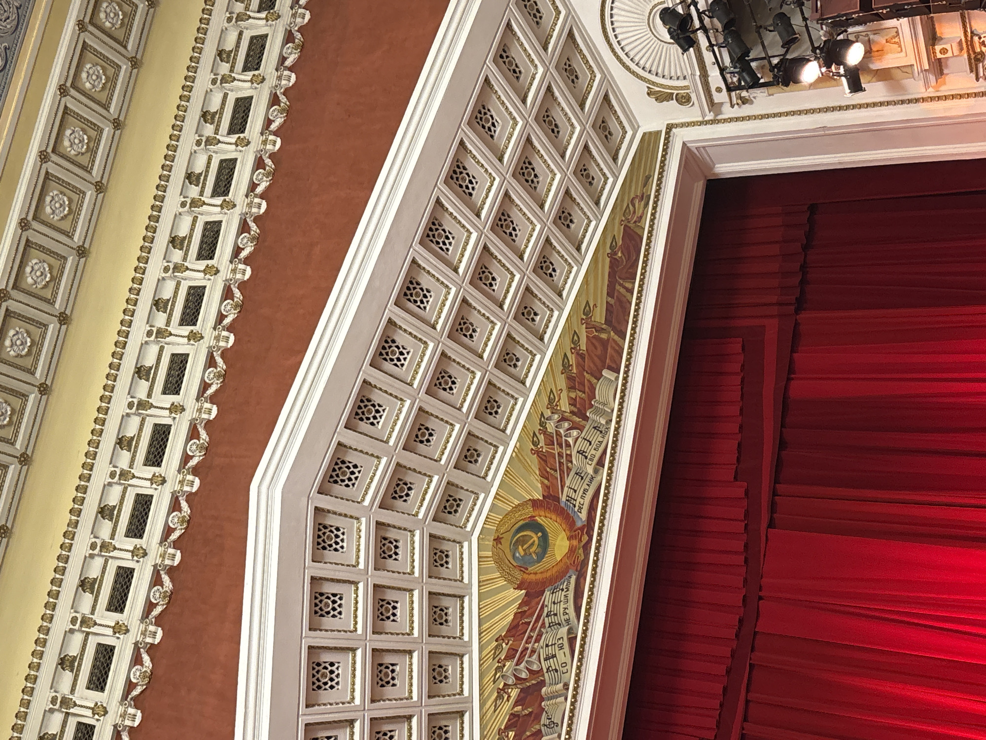 The image captures the interior of the Perm Opera and Ballet Theatre in Perm, Russia. The focus is on the ornate ceiling and the red stage curtain. The ceiling features intricate, geometric patterns and a mural containing Soviet imagery, including the hammer and sickle emblem. Stage lighting fixtures are visible near the top of the frame. The colors are primarily white, gold, red, and brown.