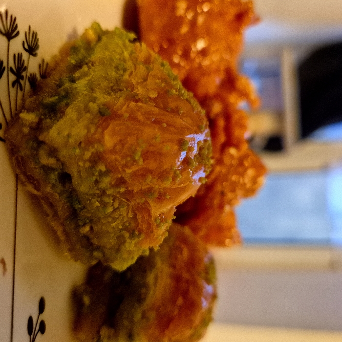 A close-up captures two pieces of baklava, one featuring a green nut topping and the other glazed in a shimmering orange hue, presented on a patterned plate with black botanical art. The pastry displays a flaky texture, glistening with syrup. Background shows blurred window.