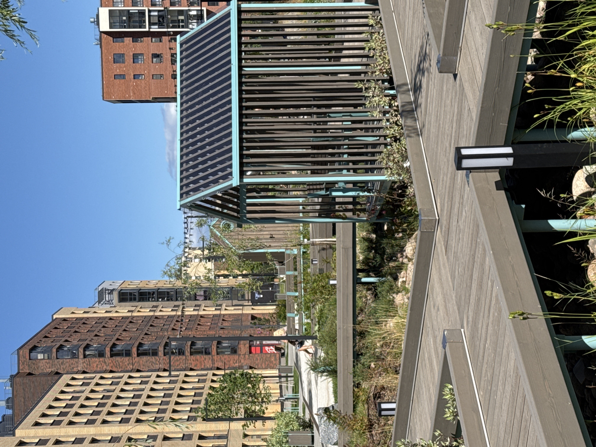 A sunny day with clear blue skies over a modern residential complex in Novoselye, Russia. The foreground features a wooden boardwalk winding through a landscaped area with tall grasses and decorative plants. Several modern, dark square lanterns are mounted on the boardwalk's railing. In the midground, a dark wooden gazebo with a slatted roof and light blue trim stands over a shallow water feature with aquatic plants. Behind the gazebo, a woman with blonde hair is seated on a light blue park bench, appearing to stretch or exercise. Further in the background, tall, multi-story apartment buildings constructed with brick and modern materials dominate the skyline. Trees and other landscaping are visible between the buildings.