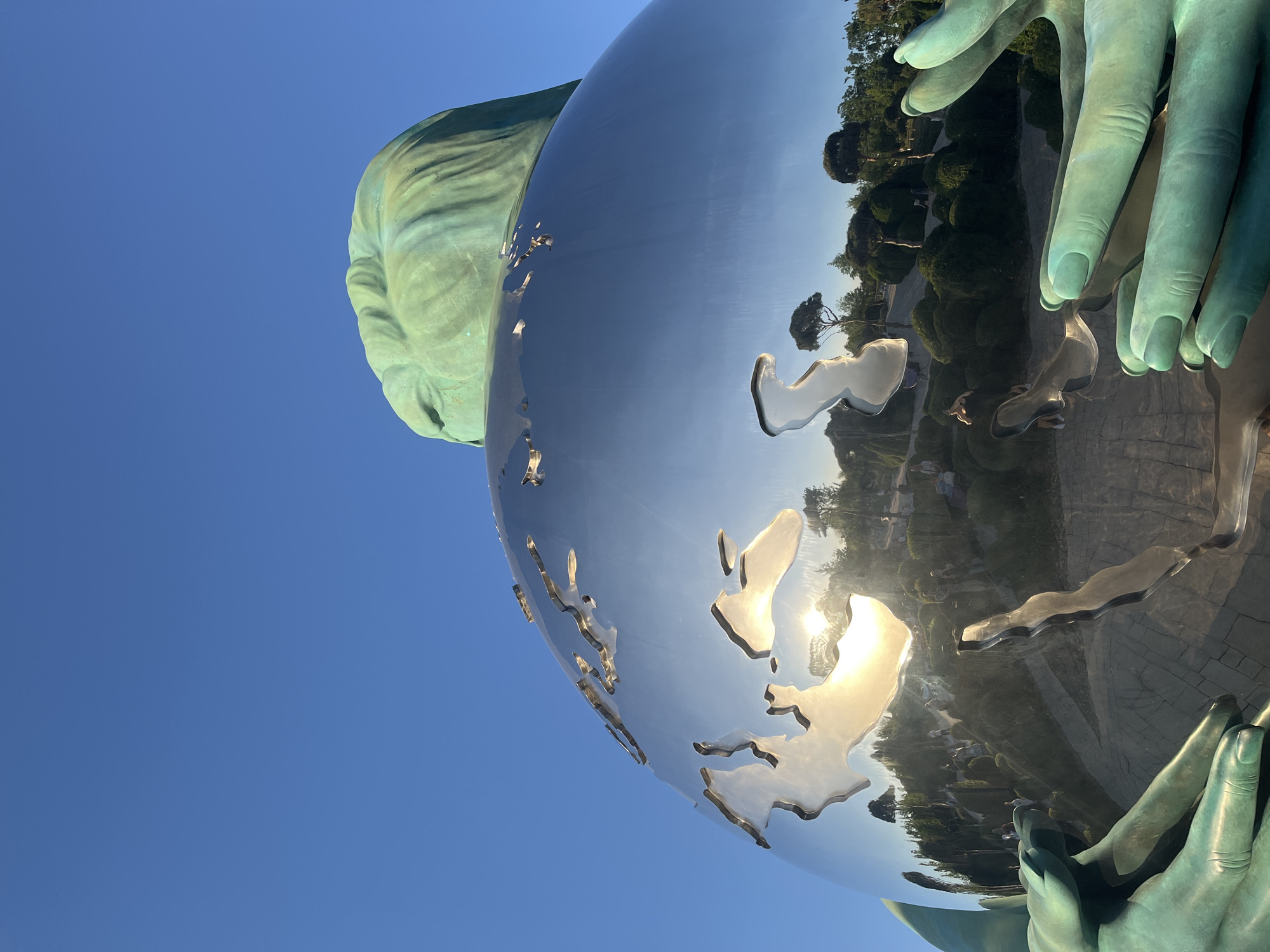 The image features a striking monumental sculpture composed of weathered bronze hands cradling a large, highly reflective metallic globe. The hands, exhibiting a greenish-blue patina characteristic of aged bronze, appear to be part of a larger figure, with a section of a head or shoulder visible on the upper left, crafted from the same material.
The globe is smooth, shiny, and silver-toned, with raised, abstract representations of continents. Its polished surface acts as a mirror, reflecting the surroundings. Prominently reflected on the globe are lush green foliage, including trees and bushes, suggesting an outdoor setting like a park or garden. A bright sunburst, indicating direct and intense sunlight, is visible on the globe's surface. The background consists of a clear, deep blue sky. Other reflections on the globe hint at paved ground or pathways, and potentially distant structures, further defining the public, open-air environment.
The scene takes place on a bright, clear day, likely late morning or early afternoon, given the intensity of the sunlight. No specific activity or event is occurring, as the image focuses solely on the static sculpture. There is no visible text in the photograph. The sculpture, located in Krasnodar, Russia, powerfully depicts hands supporting the Earth, evoking themes of environmental stewardship, global connection, or responsibility.