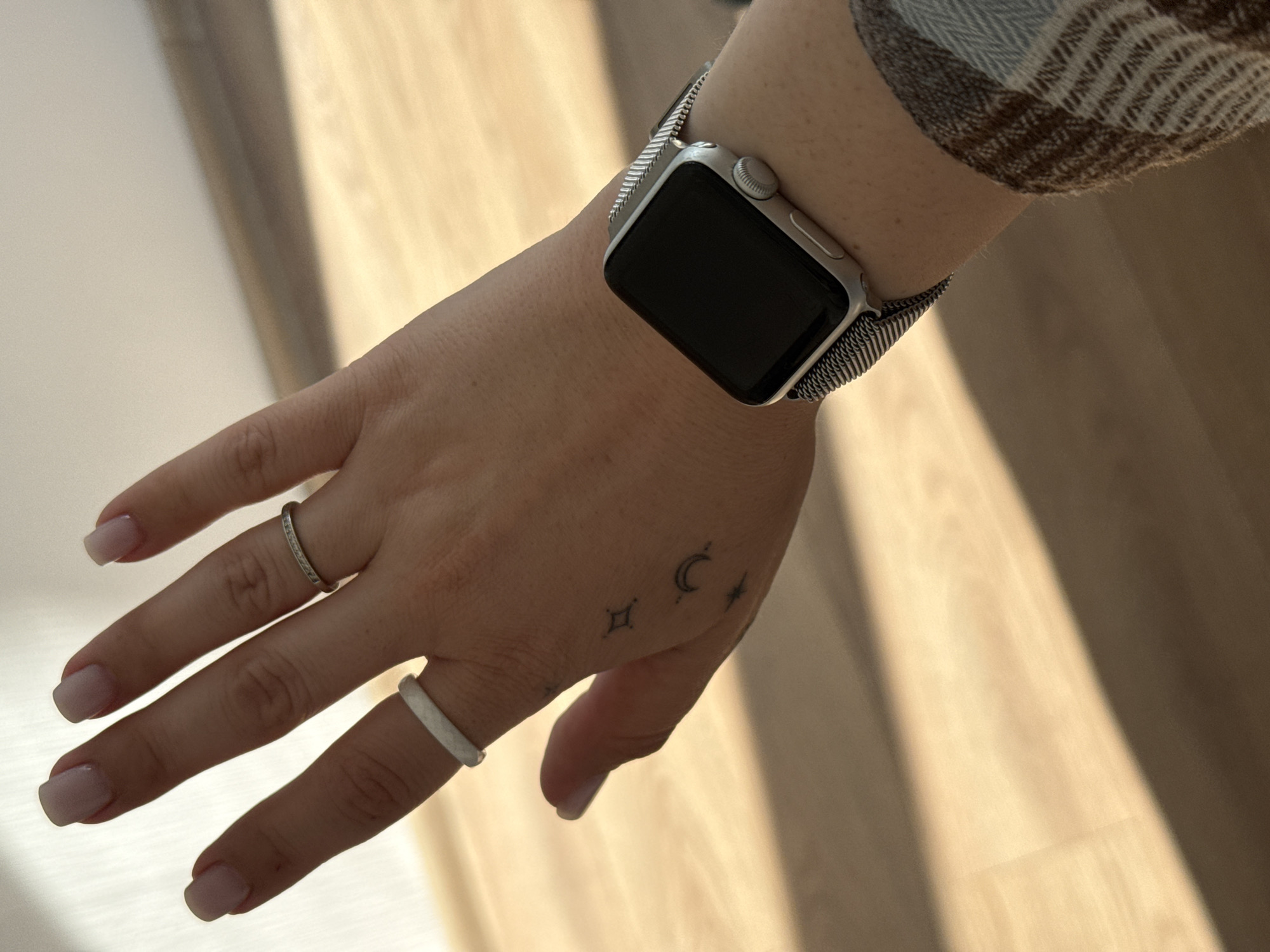 A person's left hand, adorned with two rings and a smartwatch, is positioned in front of a light-colored, wood-paneled wall, illuminated by diagonal light. The hand exhibits light-toned fingernails and a small tattoo of celestial symbols on the back. The smartwatch, a silver Apple Watch with a mesh band, is worn on the wrist. The overall scene is one of personal adornment and a dimly lit indoor environment, with no discernible urban features linking it to Lyubertsy, Russia.
