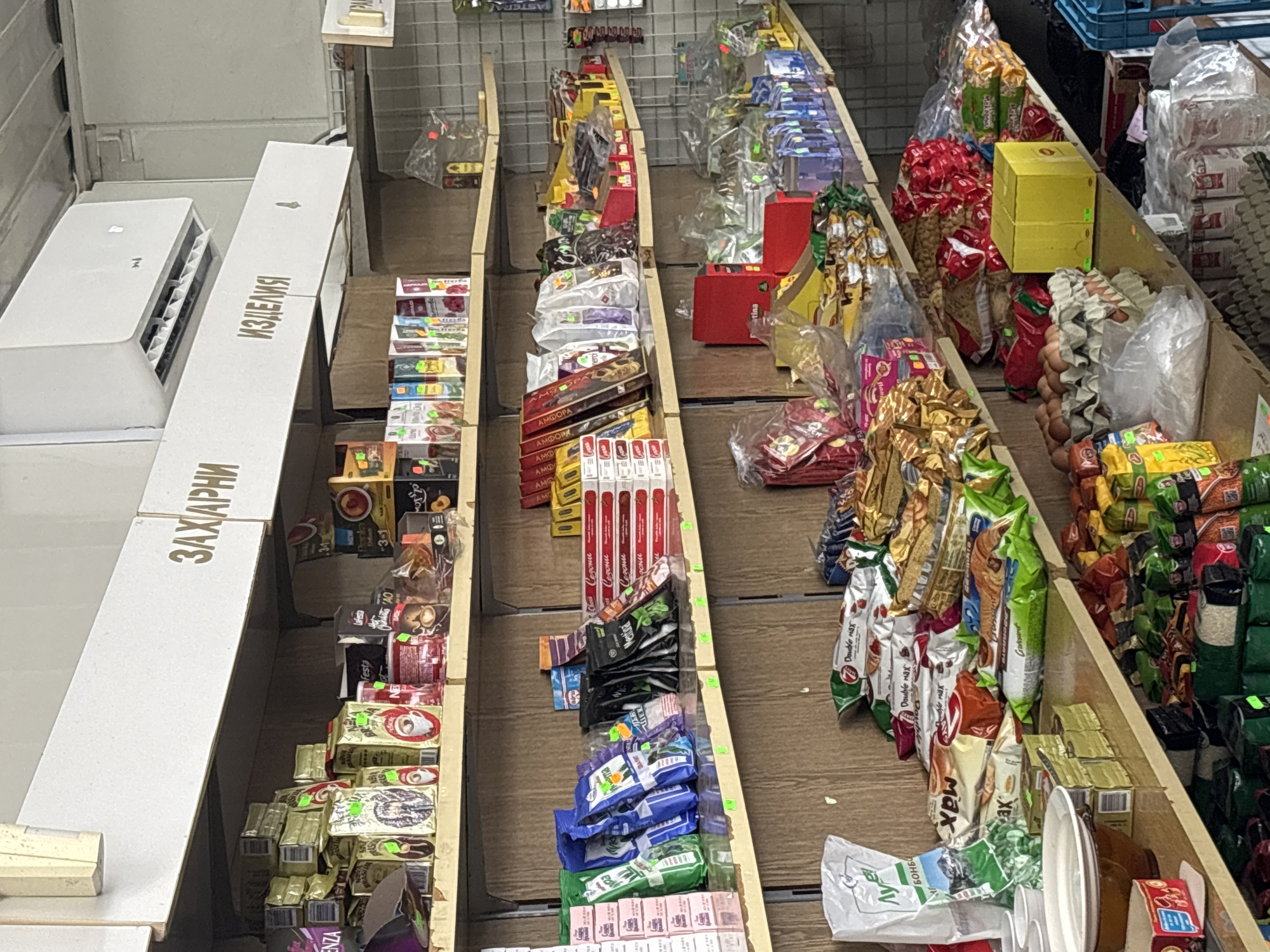 This image depicts a retail setting, likely a small grocery store or convenience store in Gurkovo, Bulgaria, possibly a market stall. The primary focus is on shelves stocked with various food products and packaged goods, creating a sense of abundance and variety.
The scene is characterized by an array of packaged items, including what appear to be sweets, chocolates, biscuits, coffee, and other snacks. Prominently displayed are the text "ИЗДЕЛИЯ" (products) and "ЗАХАРНИ" (confectionery) on white signage, suggesting a focus on sweet treats. Many packages feature colorful designs and some Cyrillic text, such as "Festa," "AMO," "Double max," and "Croissant."
The arrangement of goods on wooden shelves suggests a somewhat cluttered but organized display. Some items are packaged in plastic bags, while others are in cardboard boxes or wrappers. The presence of an air conditioner unit on the left side of the image provides a clue to the indoor setting, and the lighting suggests it could be daytime, though the exact time is not discernible. The overall impression is one of a functional retail space where goods are presented for sale. There are no people visible in the image. The scene is static, focusing on the merchandise itself.