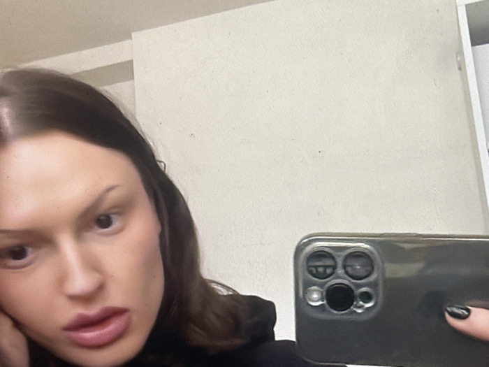An indoor scene captured as a mirror selfie. A female individual with long, dark hair, dark eyes, and full lips is partially visible on the left, looking directly towards the viewer's perspective. Her left hand is positioned near her chin. Her right hand, with black nail polish visible on the thumb and forefinger, holds a dark-cased smartphone (identifiable by its triple-lens camera array) to capture the image. The background is a plain, light-colored interior wall, possibly off-white, with a subtle texture and faint vertical seams. A narrow vertical white element is present on the extreme right, suggesting a doorframe or cabinet edge. The specific location within Tunbridge Wells, United Kingdom, cannot be determined from the visual evidence provided.