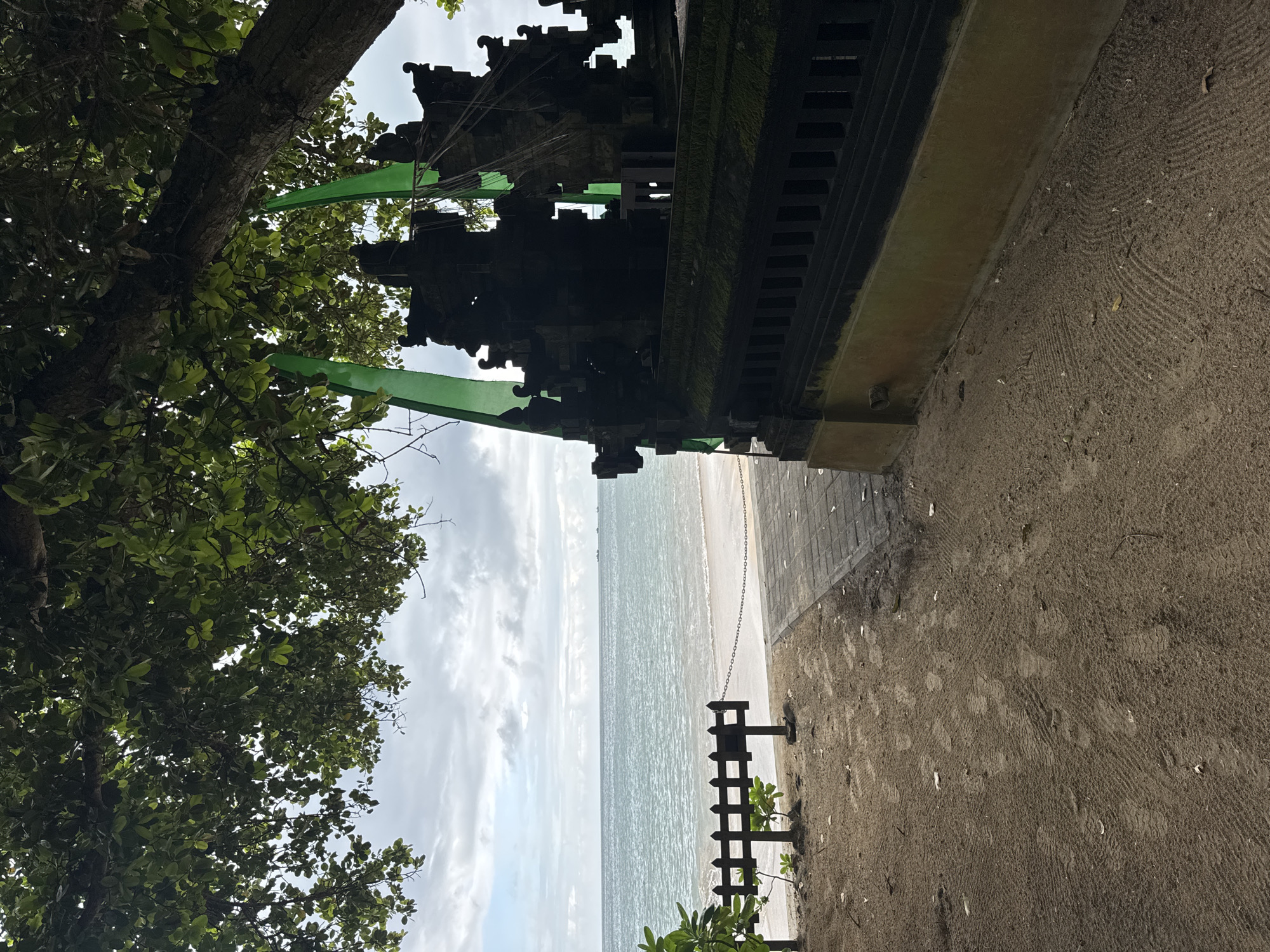 This image presents a serene coastal landscape, likely in Kuta Selatan, Indonesia, viewed under an overcast sky.
The scene is dominated by a harmonious blend of nature and traditional architecture. On the left, a large tree with lush green foliage fills a significant portion of the frame, its dense canopy and visible branches suggesting a mature tree. To the right of the tree, a dark, ornate stone structure, possibly a Balinese temple gate or a traditional building, stands prominently. This structure features intricate carvings and a weathered texture, with some moss or discoloration on its surfaces. Green banners or fabric strips are draped near the tree and the upper part of the structure, adding a subtle touch of color.
Beyond the architectural element, a wide expanse of calm, light blue-grey ocean stretches towards the horizon, meeting a partly cloudy sky. A narrow strip of light-colored sandy beach runs along the coastline, separating the land from the water. In the lower right foreground, a small, dark wooden fence or barrier is partially visible on the sand, with a metal chain attached. The immediate foreground consists of sandy or dirt ground, showing distinctive raked patterns or foot imprints, and a paved area adjacent to the stone building.
The image captures a tranquil daytime moment with diffused light, indicating a non-sunny yet bright day. The combination of the traditional stone architecture, tropical vegetation, and the ocean view strongly suggests a location rich in cultural heritage and natural beauty, consistent with the provided context of Bali. No people or visible text are present, enhancing the peaceful and timeless quality of the scene.