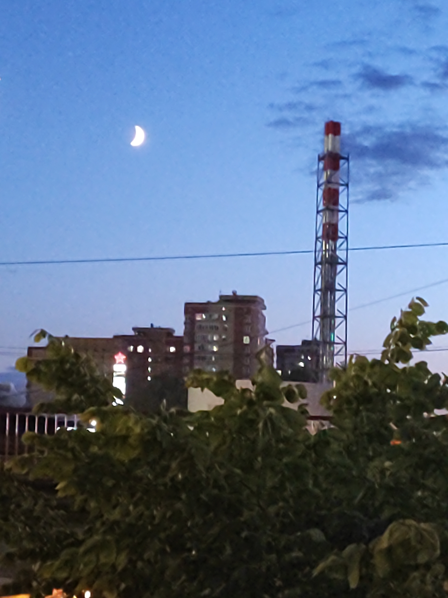 In a cityscape at dusk, a slender crescent moon hangs in the clear, deep blue sky, casting a soft glow. A tall, industrial smokestack, striped red and white, stands as a prominent vertical element against the horizon. In the mid-ground, a row of apartment buildings are visible, their windows lit, suggesting an urban residential area. A bright red star emblem is illuminated on one of the buildings. The foreground is dominated by the lush, dark green foliage of trees, their leaves obscuring the lower portions of the scene. A thin power line runs horizontally across the frame, dividing the sky and the cityscape. The overall impression is a blend of natural celestial beauty and the man-made structures of an urban environment during twilight.