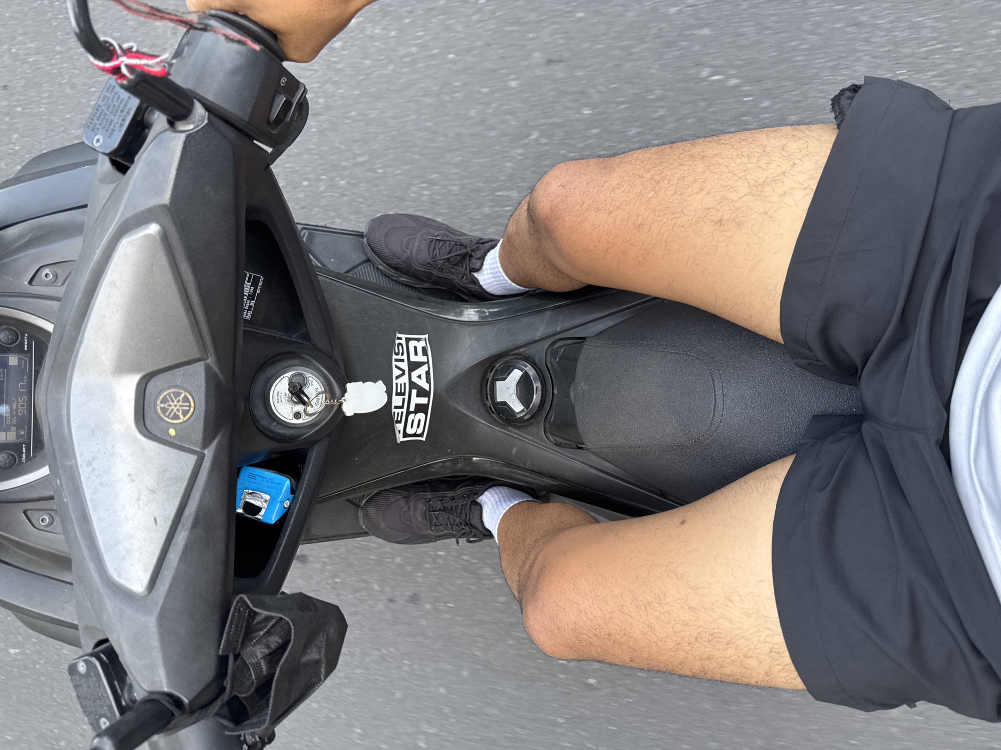 The image is a first-person perspective shot of a person riding a dark-colored scooter, likely in Kuta Selatan, Indonesia, given the context. The rider is wearing black shorts and black athletic shoes with white socks. The scooter's dashboard is visible, showing a digital display and ignition. A black face mask is hanging from the left handlebar, and a small blue object is tucked into a storage compartment. The road surface appears to be asphalt, and the lighting suggests it is daytime. A sticker with the text "ELEVISTAR" is visible on the scooter's body. The rider's legs are positioned on the floorboard of the scooter.