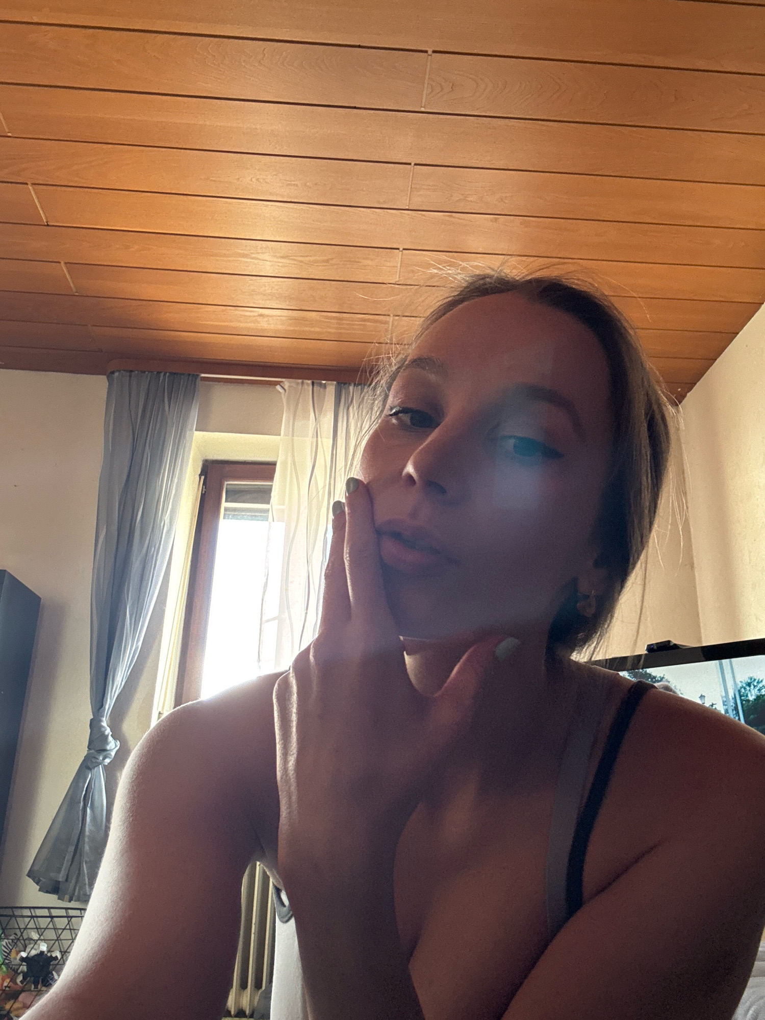 The image shows a young woman indoors, with her face partially obscured by her hand. She is looking towards the camera, and her pose suggests a selfie. The background includes a window with curtains, a wooden ceiling, and a television screen. The lighting is dim, with natural light coming from the window. The scene appears to be a bedroom or living area. There is no specific indication of the location being in the city of Überherrn, Germany.
