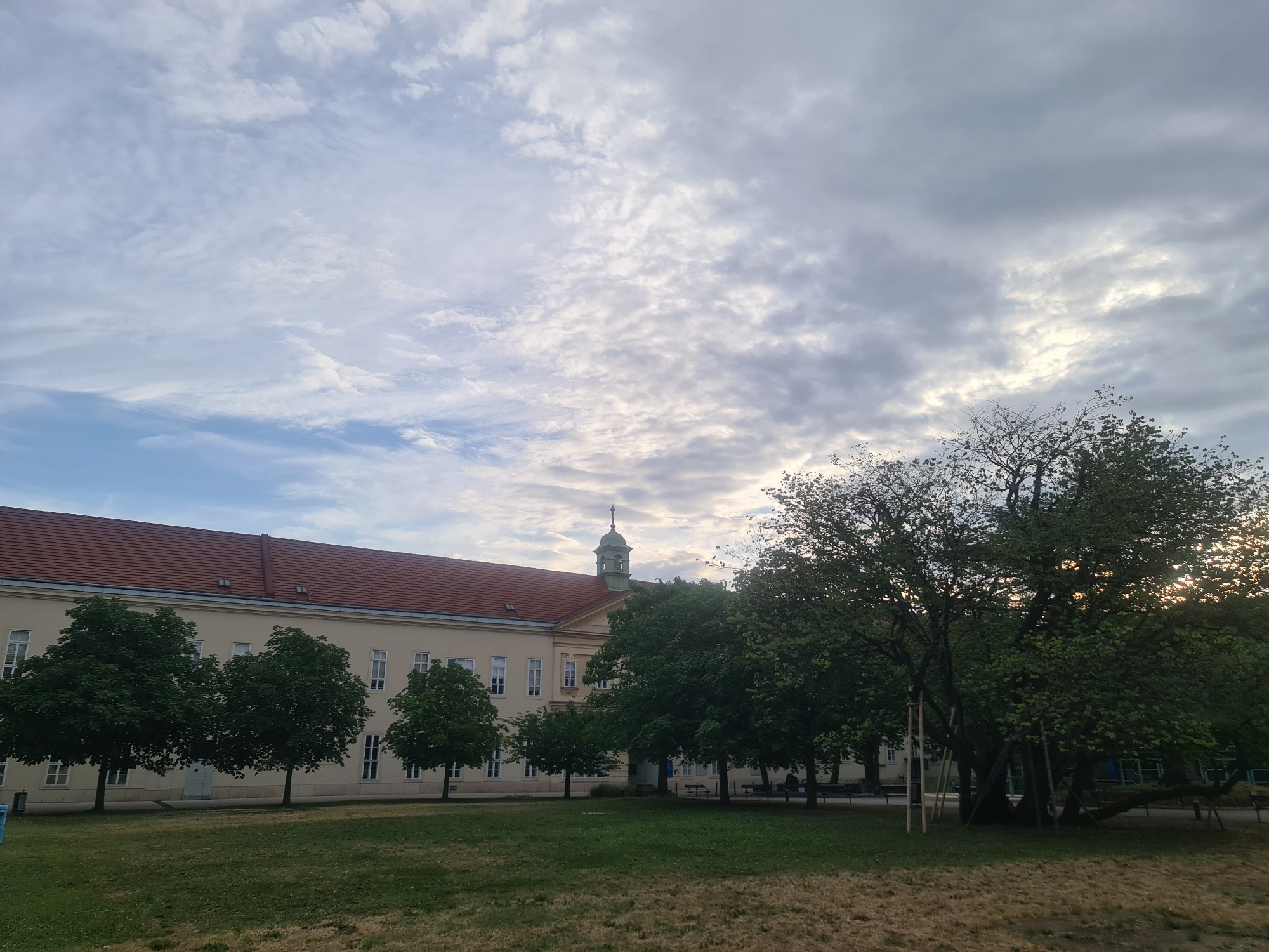 The image depicts a building with a red tiled roof and pale yellow walls, featuring a central tower with a green spire. The building appears to be an institutional or educational facility, possibly a school or administrative building, with numerous windows and a symmetrical facade. In front of the building, a row of mature trees with lush green foliage is visible. To the right of the building, a large, old tree with a thick trunk and branching limbs dominates the foreground, supported by wooden stakes. A grassy lawn stretches across the bottom of the image, transitioning to a more sparse, dry grassy area towards the front. The sky is filled with a dramatic display of clouds, ranging from bright white to darker grays, suggesting either an overcast day or the aftermath of a storm. Sunlight filters through gaps in the clouds, casting a soft light on the scene. The overall impression is one of a serene, somewhat aged environment. Based on architectural style and the presence of such mature trees, it is plausible this is a location like the former Sternwarte (observatory) in the Türkenschanzpark, or a similar historical building within one of Vienna's larger parks.