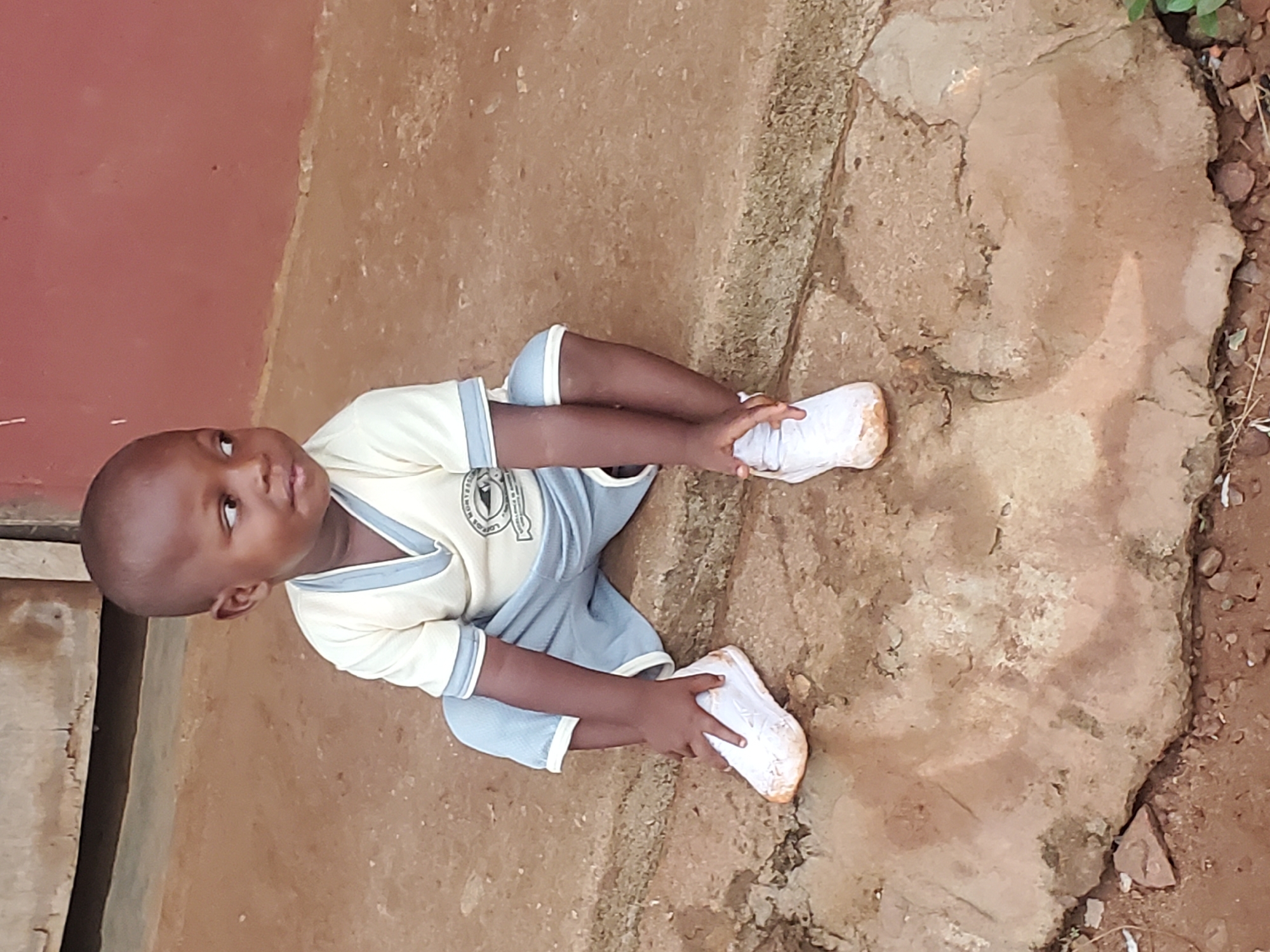 A young child is sitting on a concrete surface. The child is wearing a school uniform of light yellow and blue and has white shoes on. The child is touching their shoes. The setting appears to be outside, adjacent to a building with red paint. The location is Ukwu-Abwa, Nigeria. The ground is dirt and concrete.