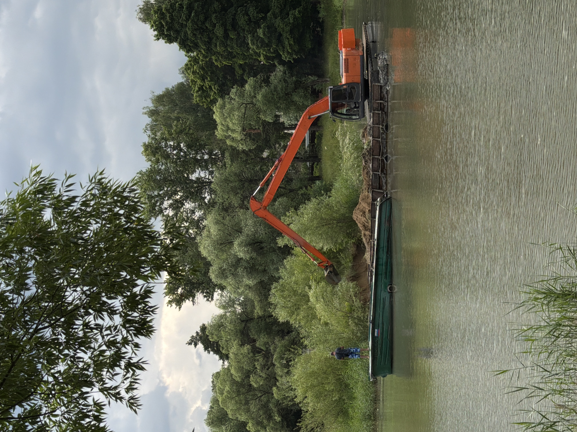 An orange excavator is dredging a river in Bykovo, Russia. The excavator is mounted on a barge and is digging sand from the riverbed, depositing it onto a separate barge. A green boat is moored next to the barges, and a person is standing on the boat. The river is surrounded by lush green trees and vegetation. The sky is overcast.