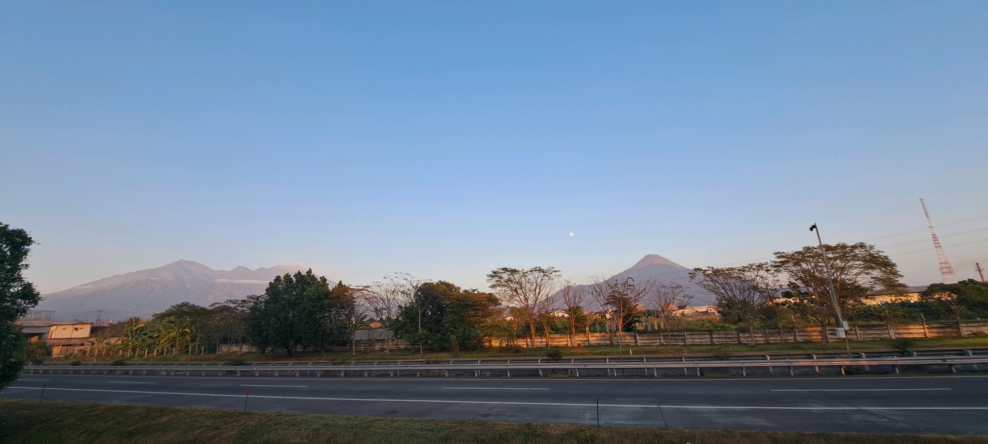 This is a wide-angle, eye-level photograph capturing a scenic view of mountains and a highway in Kebon Waris, Indonesia, during what appears to be late afternoon or early morning given the soft lighting and clear sky.
Dominating the upper portion of the image is a vast, gradient blue sky, transitioning to a lighter hue near the horizon. A small, pale circle, likely the moon, is visible in the upper right quadrant of the sky.
In the distance, a range of majestic mountains stretches across the horizon. The mountains are muted in color, suggesting they are some distance away. On the right side of the mountain range, a distinct, conical mountain peak stands out.
In the mid-ground, a highway with multiple lanes is clearly depicted. The road surface is asphalt, with visible lane markings. To the left of the highway, there's a grassy embankment. A concrete barrier separates the highway from the foreground.
Behind the highway, a line of vegetation comprising various trees and bushes forms a natural barrier. Some trees are lush and green, while others appear bare or have sparse foliage, hinting at seasonal changes or different species. A concrete fence runs behind the trees, with some buildings and structures partially visible beyond it. One building on the left appears to be a simple, light-colored dwelling with windows.
Further to the right, a tall, lattice-like communication tower with red and white sections rises against the sky. A streetlamp with a camera mounted on it stands prominently in the mid-ground, to the left of the communication tower. Power lines are also visible in the distance.
The overall scene is peaceful and serene, showcasing a blend of natural landscape and infrastructure. There are no people or animals visible in the image.