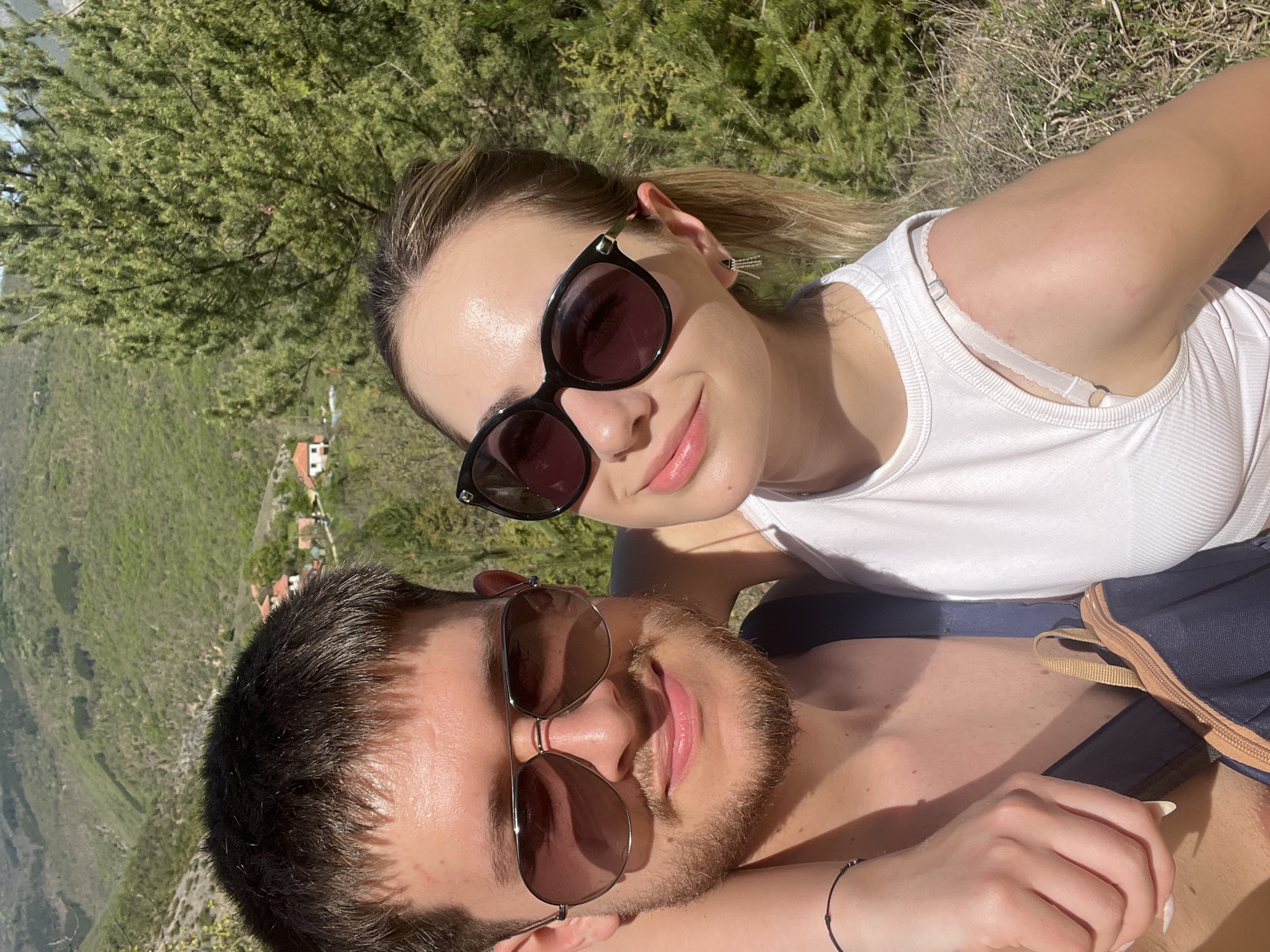 A selfie taken in a natural setting shows a woman and a man posing closely together. The woman is positioned above the man, her head slightly tilted toward the camera. She is wearing sunglasses and a white tank top. A bra strap is visible under her tank top. Her hair is pulled back in a ponytail. The man is positioned below the woman, looking slightly up at the camera. He is also wearing sunglasses. His face is partially covered with stubble and a beard. The background features green vegetation, including trees and shrubs. In the distance, there are small houses and a landscape with a valley. This location is likely near Polska Skakavitsa, Bulgaria.