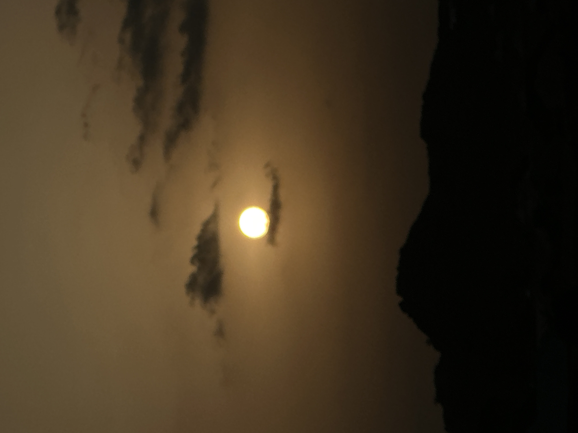 An eye-level outdoor shot shows a sun dimly visible through the clouds. The shot is taken in the city of Bukuru, Nigeria. To the right of the frame are dark rocks, as if the shot was taken with the photographer standing near a rock wall or mountain. The overall atmosphere of the shot is murky.