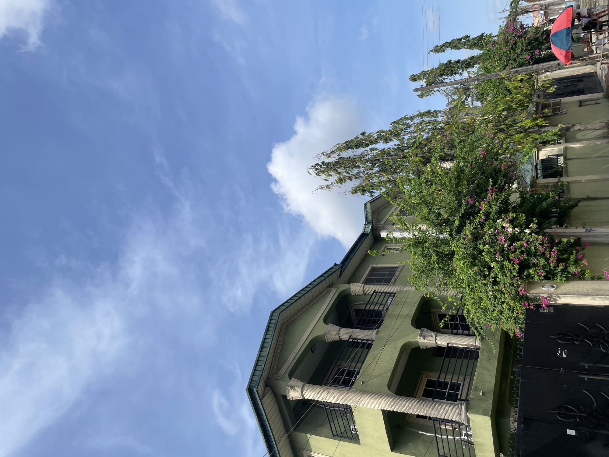 A three-story, light green building is visible in the Ado, Nigeria. It has decorative columns and black-barred windows. There are flowering plants on the side of the building, and the sky is blue with some white clouds. A small portion of a red and blue umbrella is seen on the right side of the image, along with trees.