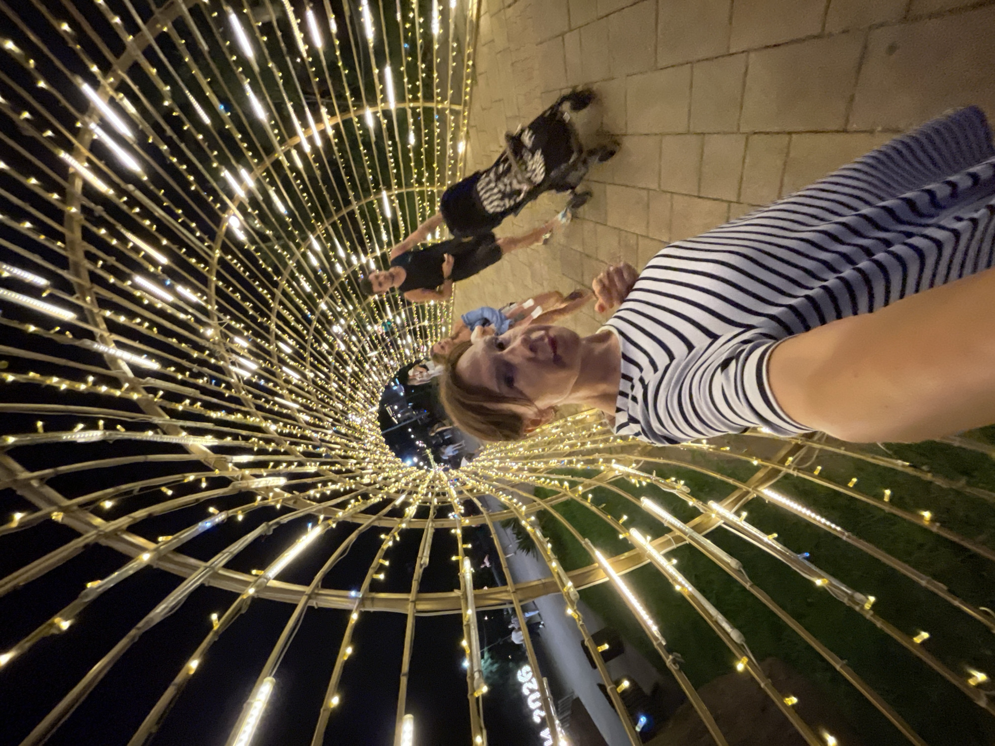 A selfie taken at night inside a brightly lit tunnel made of bamboo structures and numerous yellow string lights. The tunnel curves towards the background, creating a dramatic perspective. In the foreground, a woman with short brown hair and glasses is wearing a black and white striped shirt. She is holding the phone to take the selfie. In the mid-ground, a man in a dark tank top and shorts pushes a stroller, while a woman in a light blue top stands next to him. In the background, other people are visible walking. On the left side of the tunnel, a sign reads "2026". The ground is paved with light-colored rectangular tiles. The overall ambiance is festive and illuminated, suggesting a public gathering or attraction in Sochi, Russia.