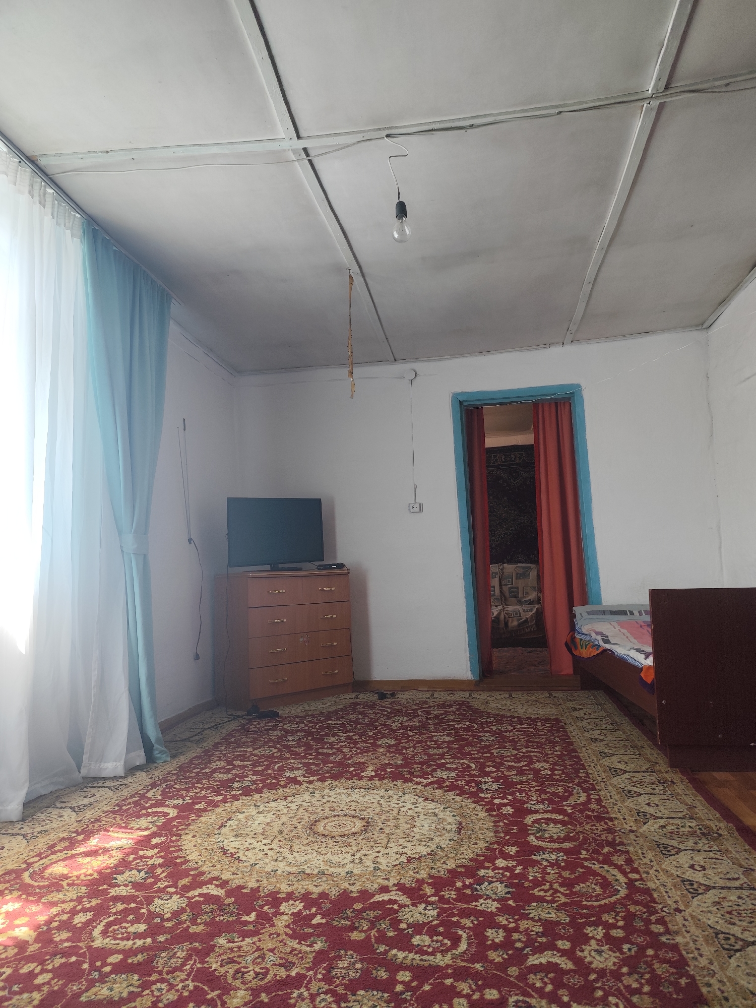 The image depicts an interior view of a room, likely a bedroom or living area, in a dwelling located in the Aqbulaq Rural District, Kazakhstan. The room is furnished with a dresser with drawers, a television set, a bed, and a large patterned rug covering the floor. The walls are painted white, and the ceiling is paneled. A lightbulb hangs from a cord in the center of the room. Curtains, one white and one light blue, are visible on the left side of the image, framing a window. An open doorway on the right side of the room reveals an adjacent space with red curtains. The overall impression is of a modest, lived-in domestic interior.