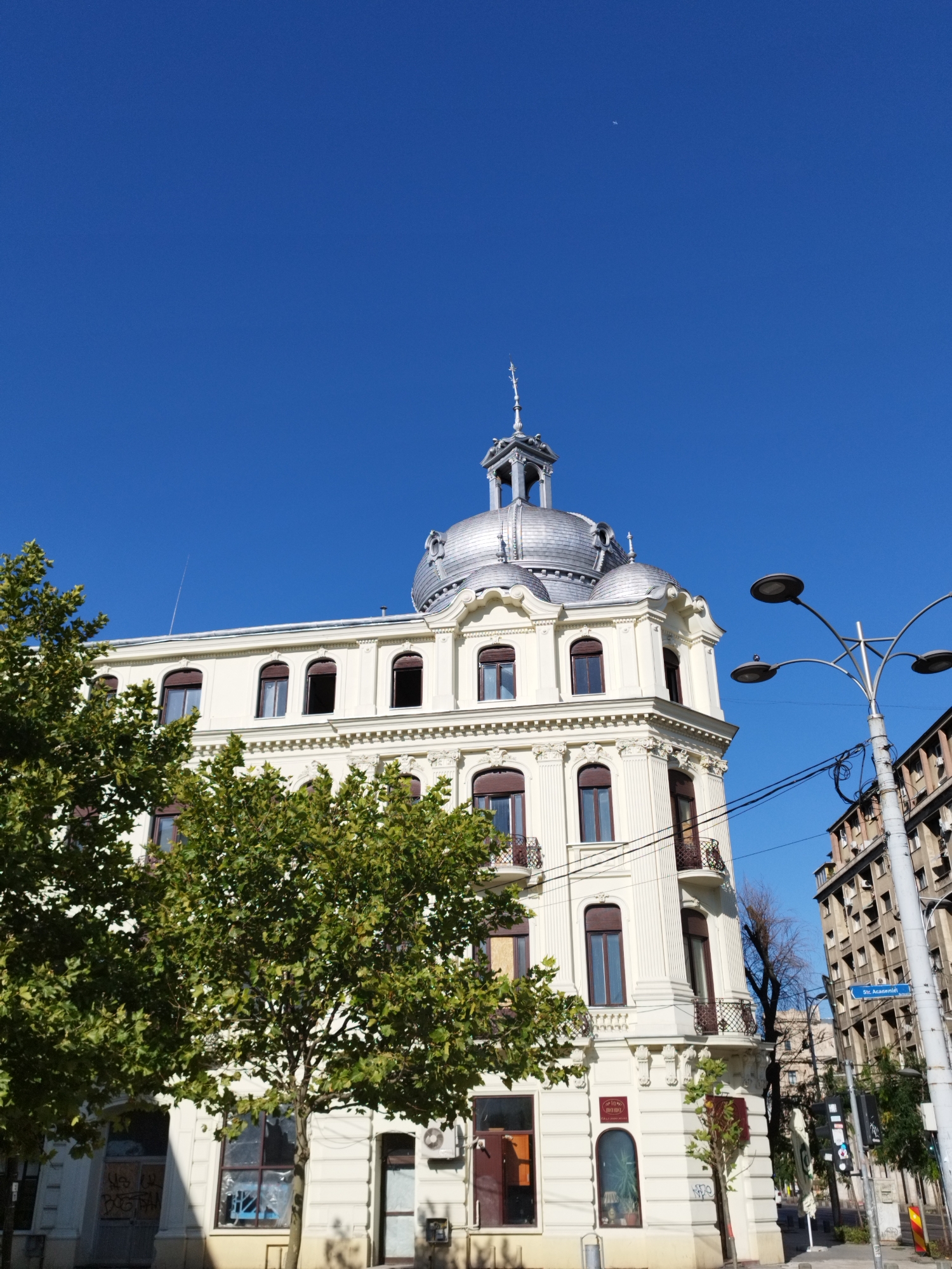The image shows a corner building with a domed roof, typical of Belle Époque architecture, located in Bucharest, Romania. The building is painted cream and has arched windows with dark brown frames. Green trees are in the foreground, obscuring part of the lower floors. A modern lamppost with multiple lights stands to the right of the building, with power lines running across the frame. In the background, another taller, older building with numerous balconies is visible. A street sign reads "Str. Acateniei," indicating the street name. The sky is clear and blue. There are no people visible in the image. The overall scene depicts an urban environment with a mix of architectural styles.